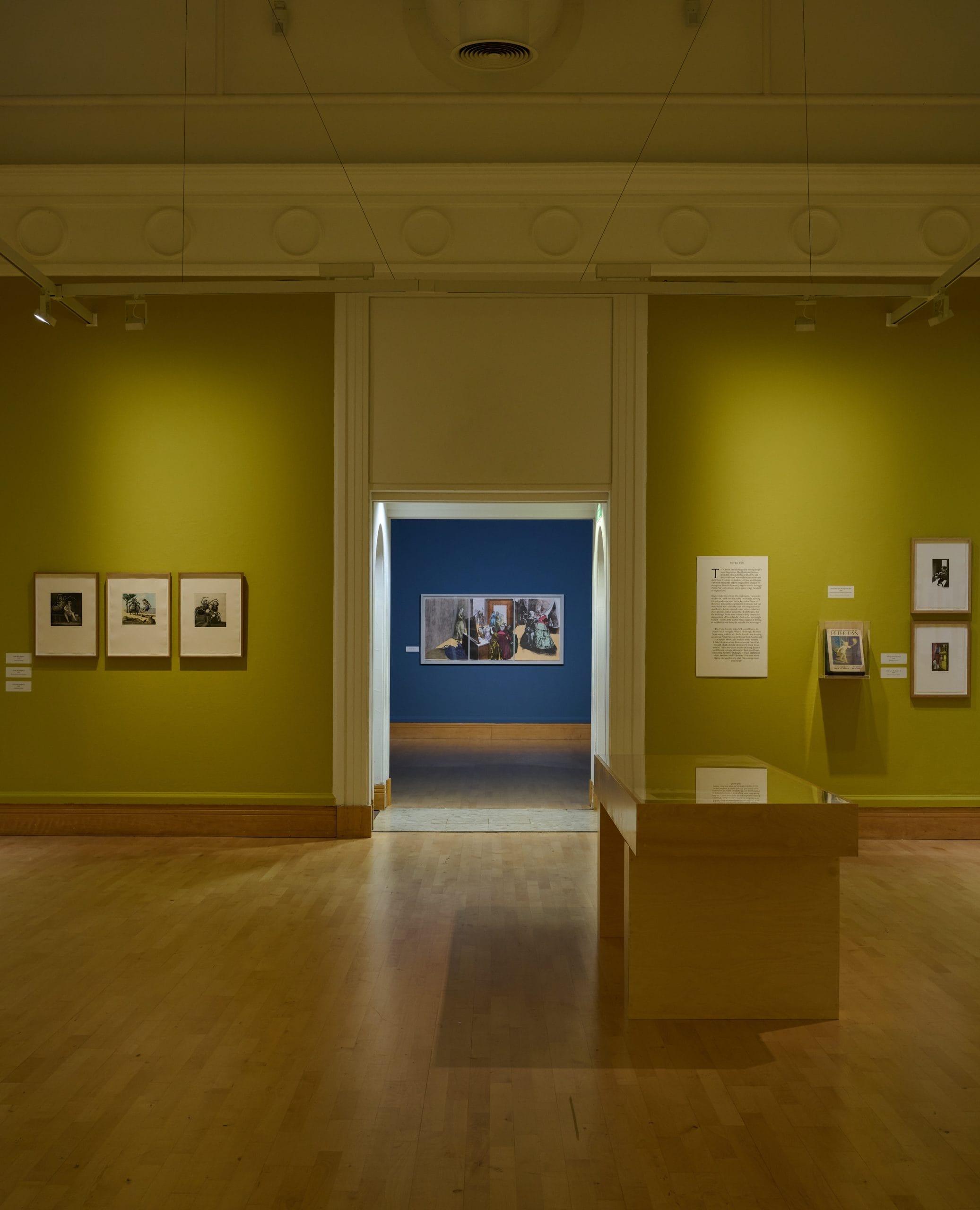 A gallery space with wooden floors and yellow walls with prints by Paula Rego on the walls.