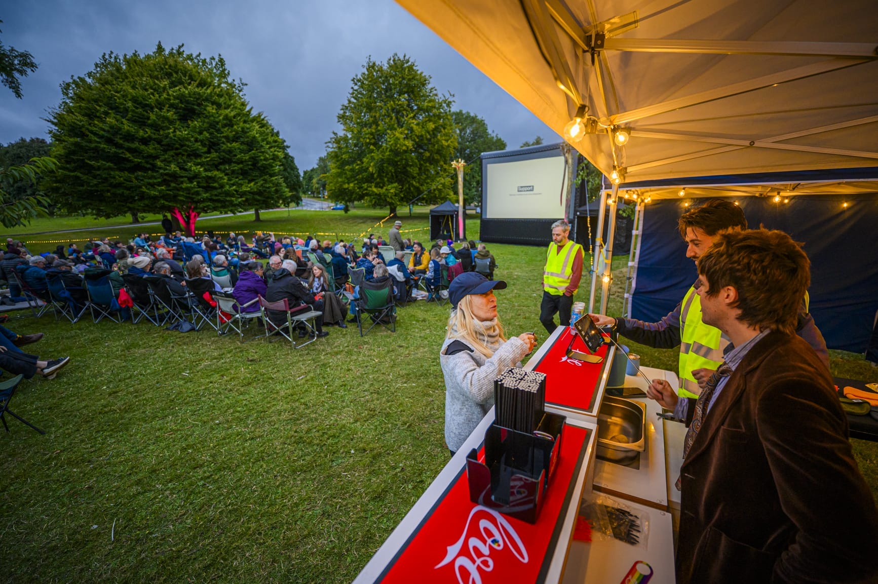 a blond woman is at the bar with open air cinema goers and the screen visible in the background