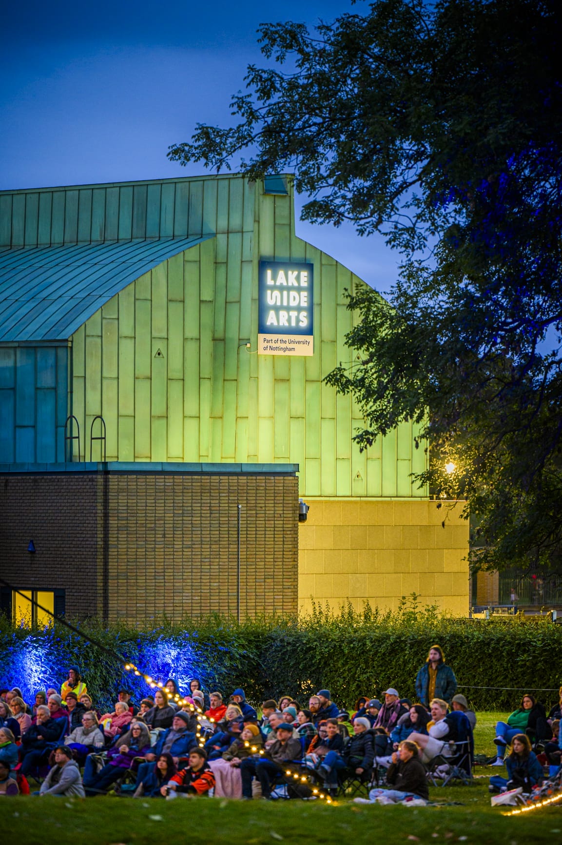 audiences watching outdoor cinema sitting on camping chairs and blankets with Lakeside's Pavilion building in the background