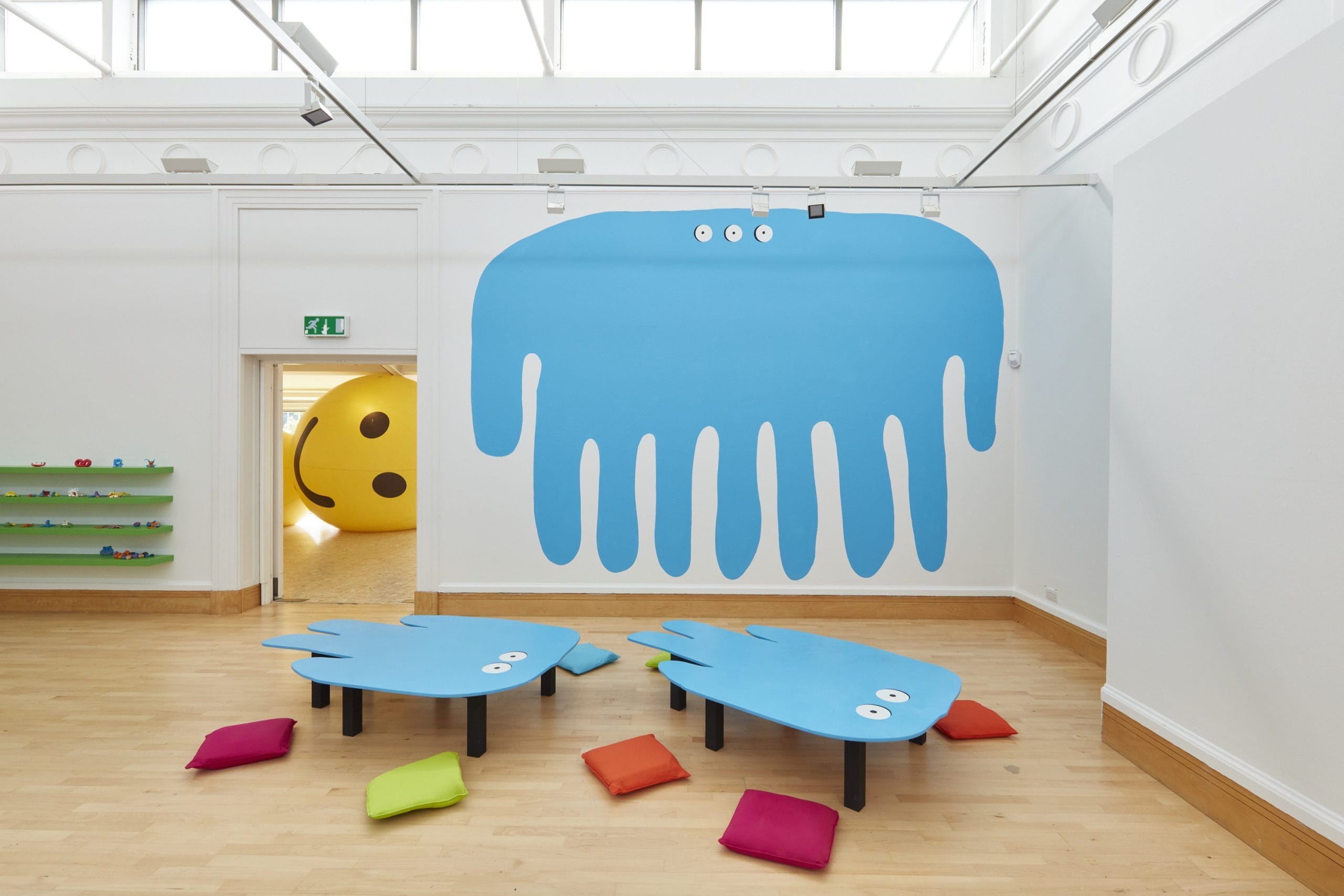 A white gallery space has a blue multi-legged monster painted on the wall and an inflatable yellow worm, with a smiley face, poking through the door.