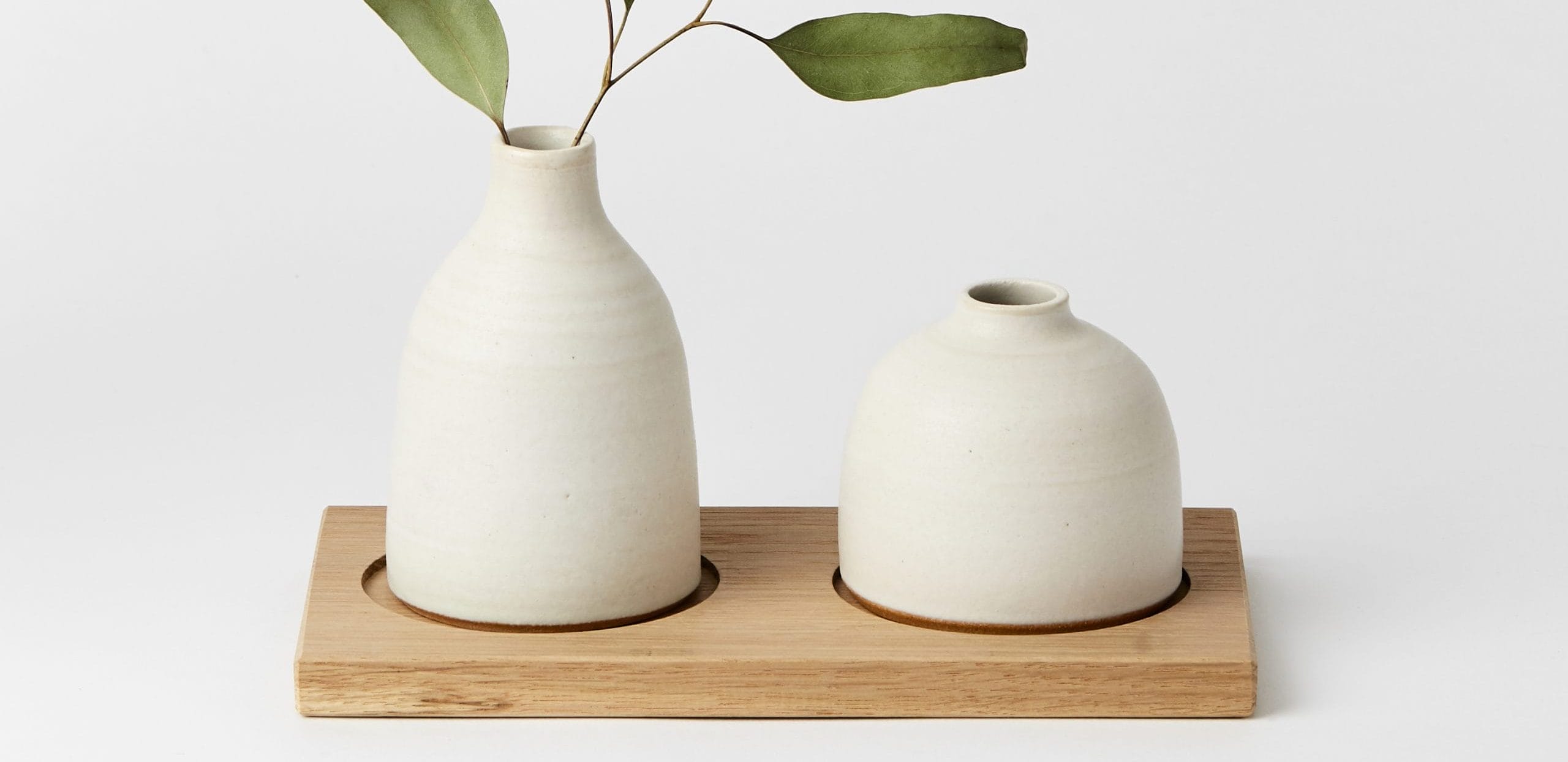 two cream vases, one with a plant stem in. They are on a wooden board.