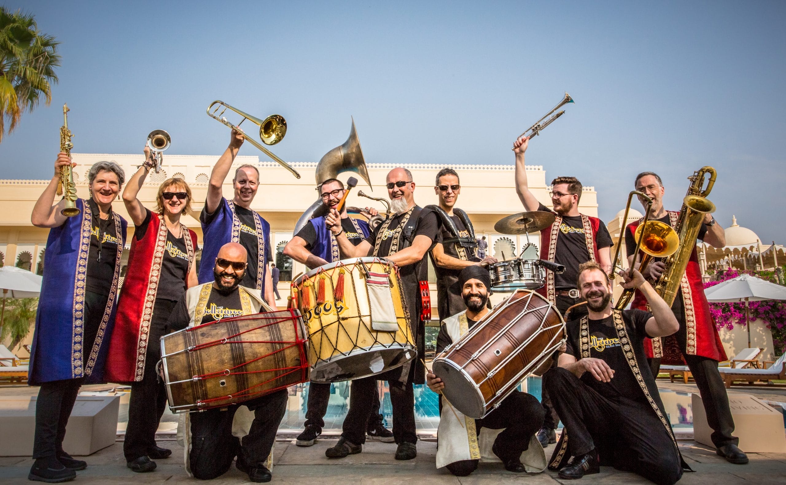 A large group of musicians, wearing long Bollywood inspired waistcoats holding drums and brass instruments.