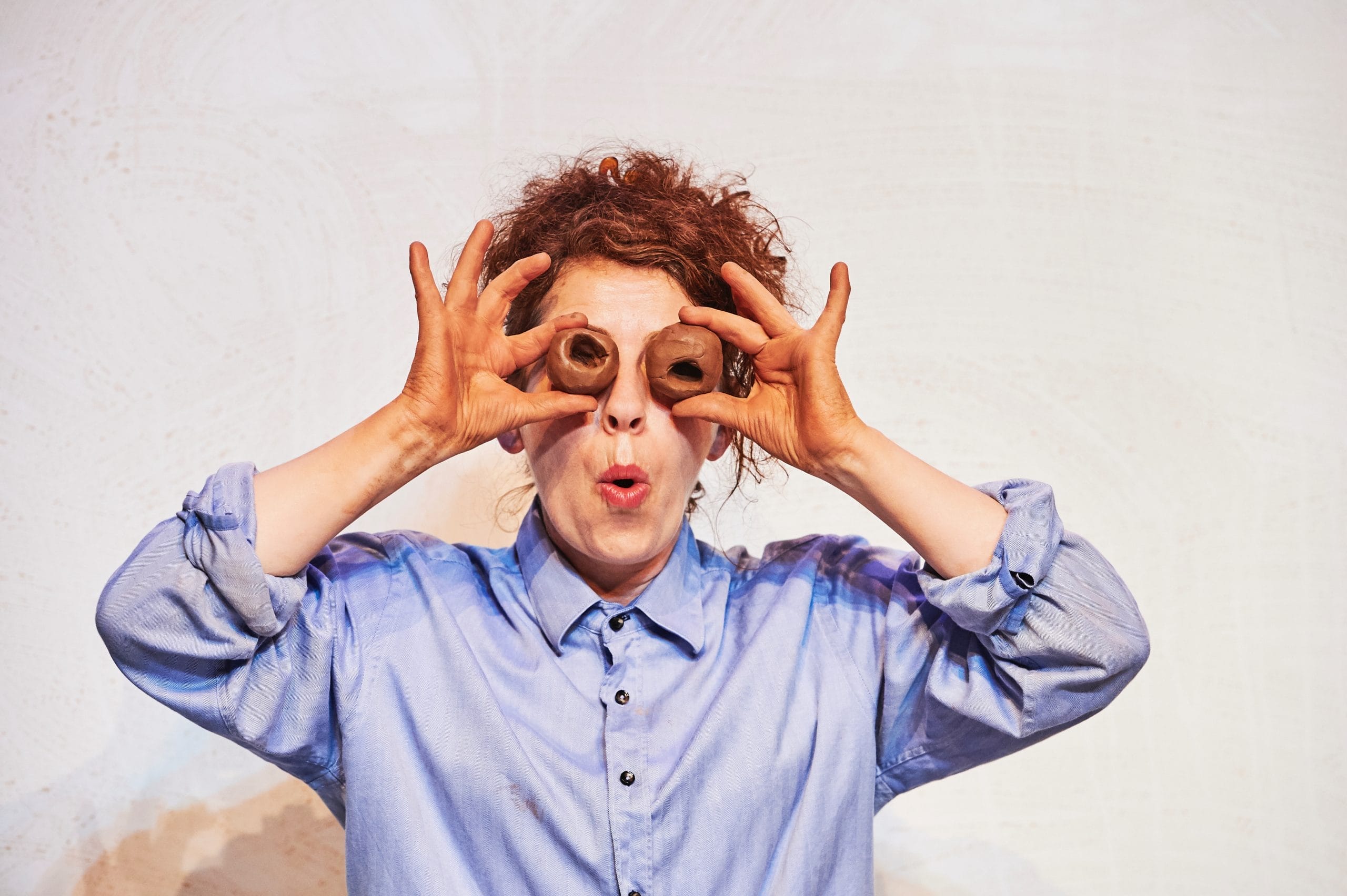 A lady in a blue shirt with curly hair, holds two clay donuts to her eyes!