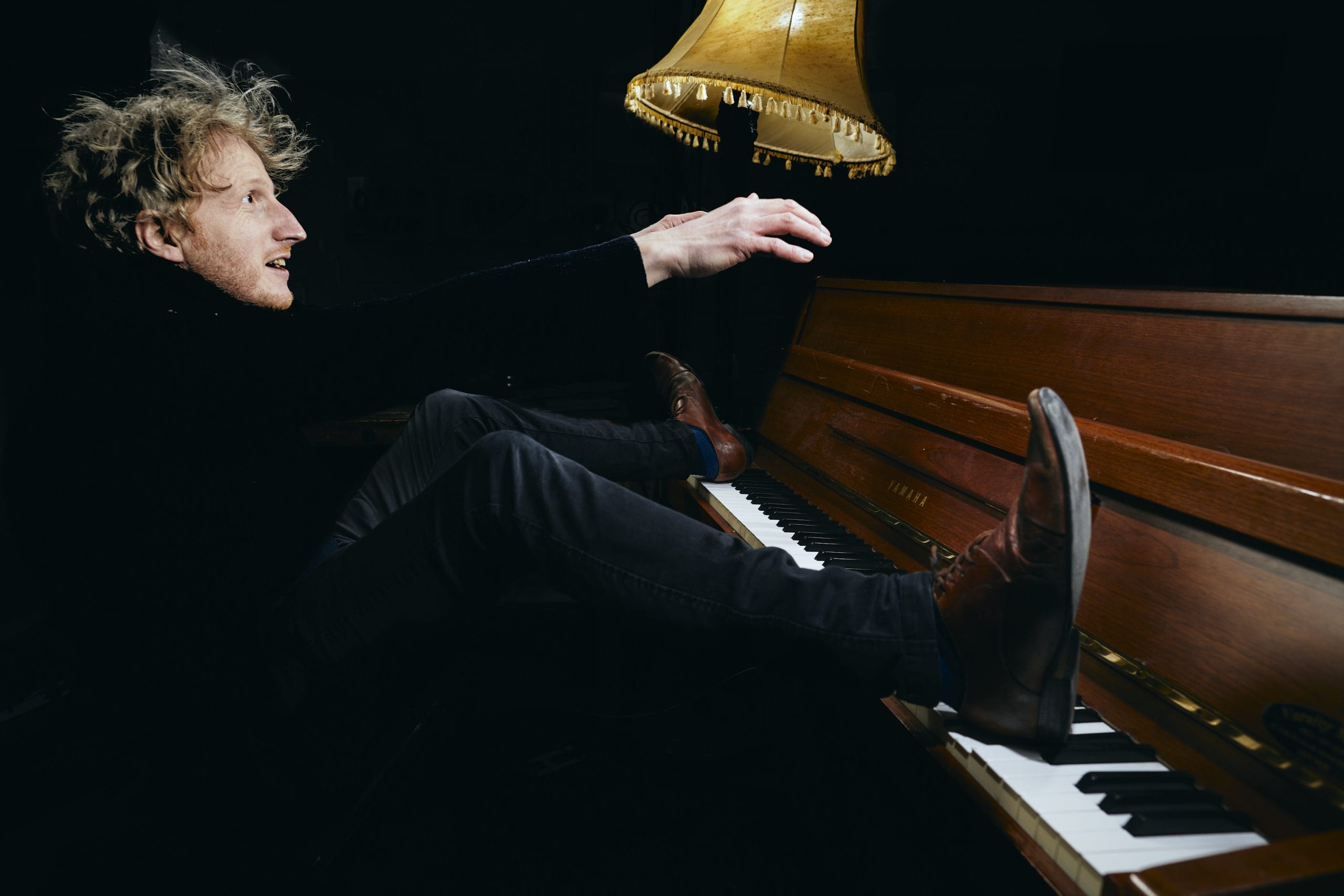 A man with crazy blond hair, dressed in dark clothes sits with his feet on a piano.