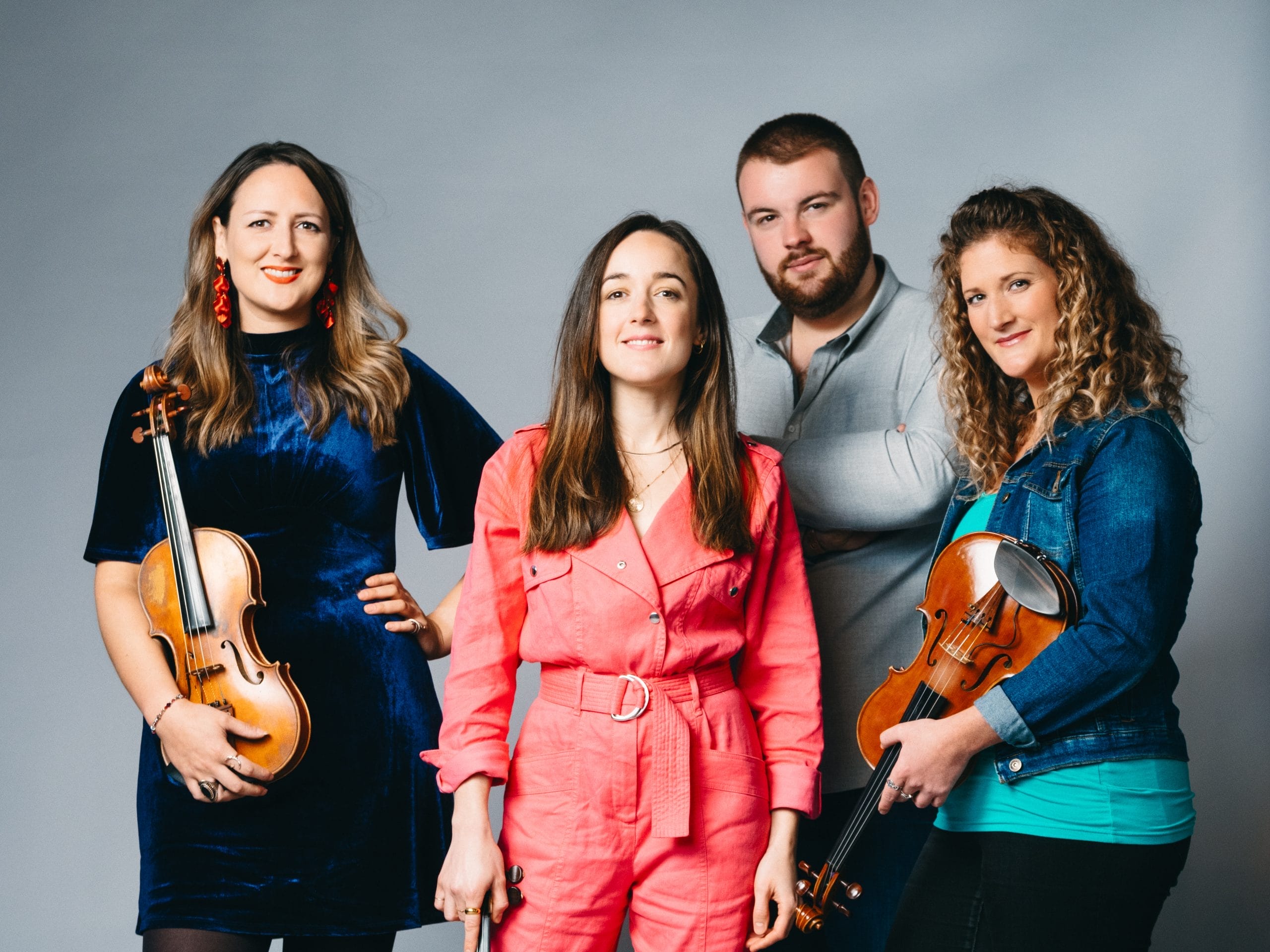 A group of musicians, three women and one man stand in front of a pale blue studio wall. All three women, wearing their hair down, are holding fiddles.