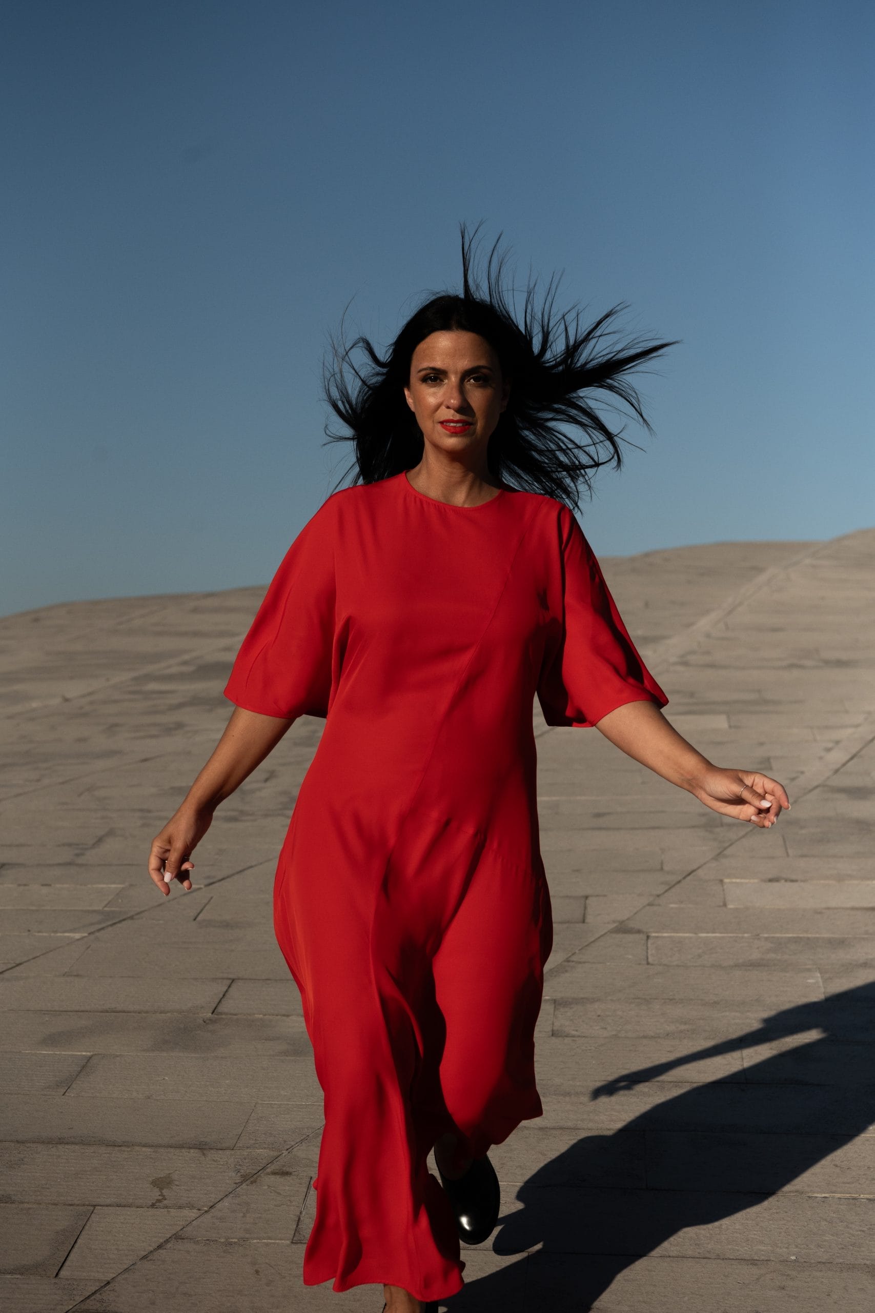 A Portuguese woman, with long flowing dark hair, wearing a red dress, walks across sand towards the camera.