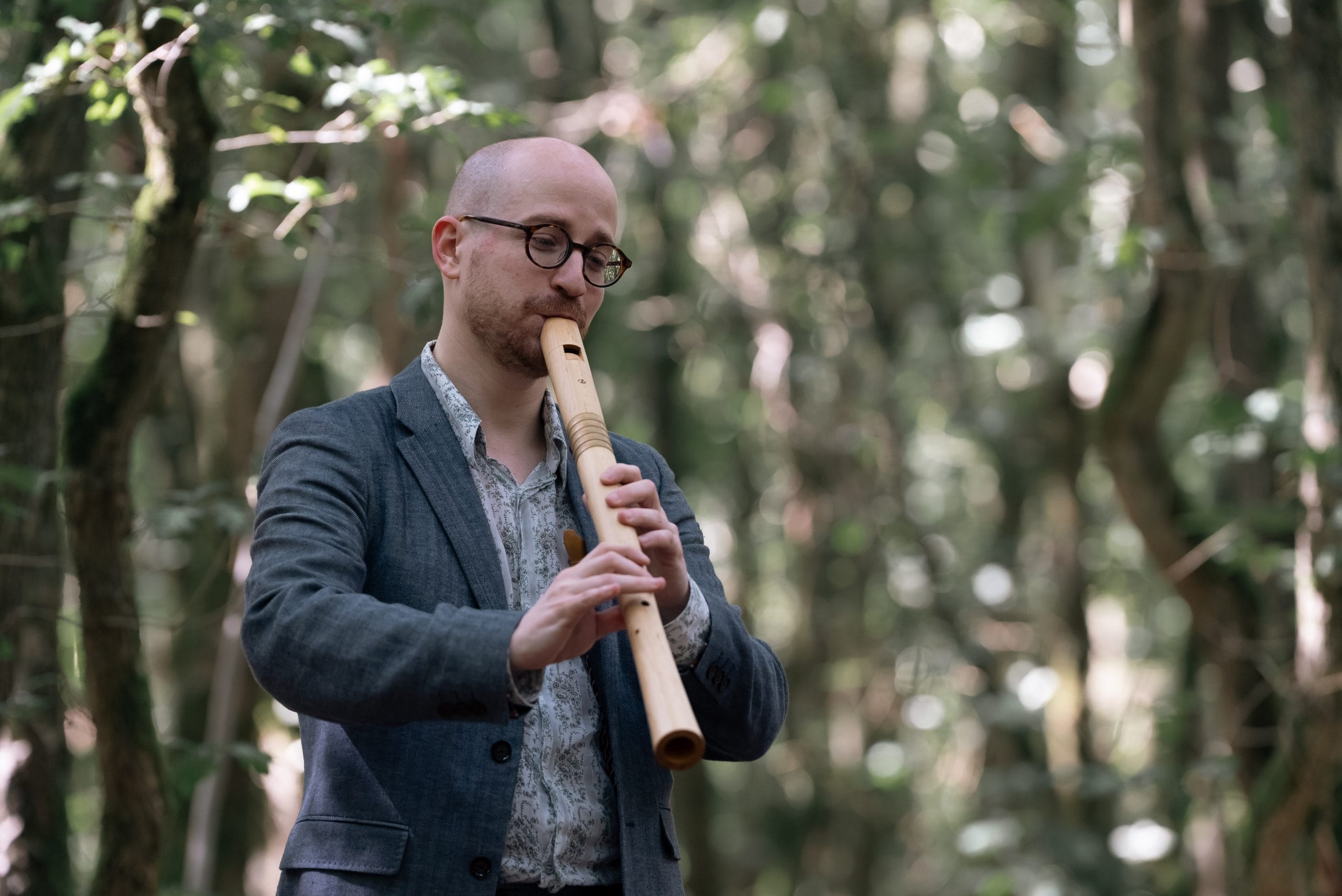 A man, with a shaved head and glasses stand amongst trees playing a larger recorder.