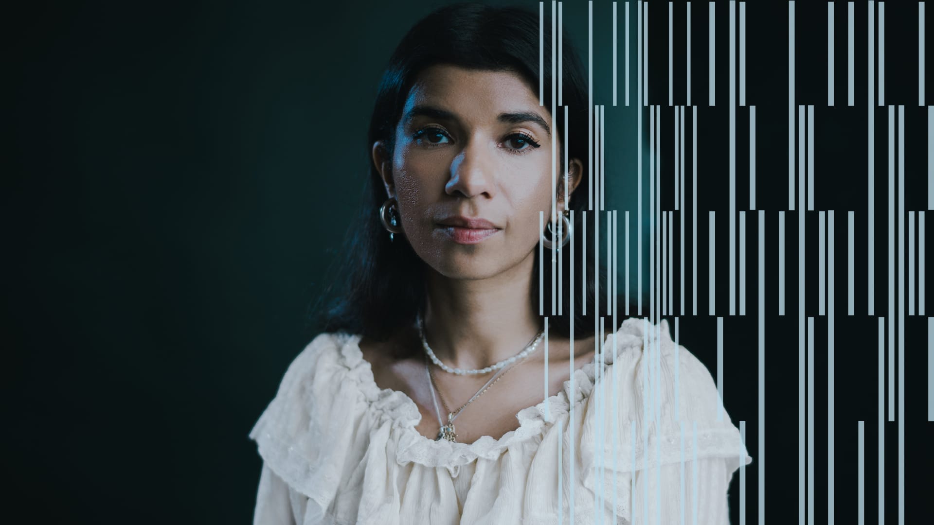 A woman with dark hair, wearing a white blouse stands in front of a black wall with a light pattern sitting over the right hand side of the image.