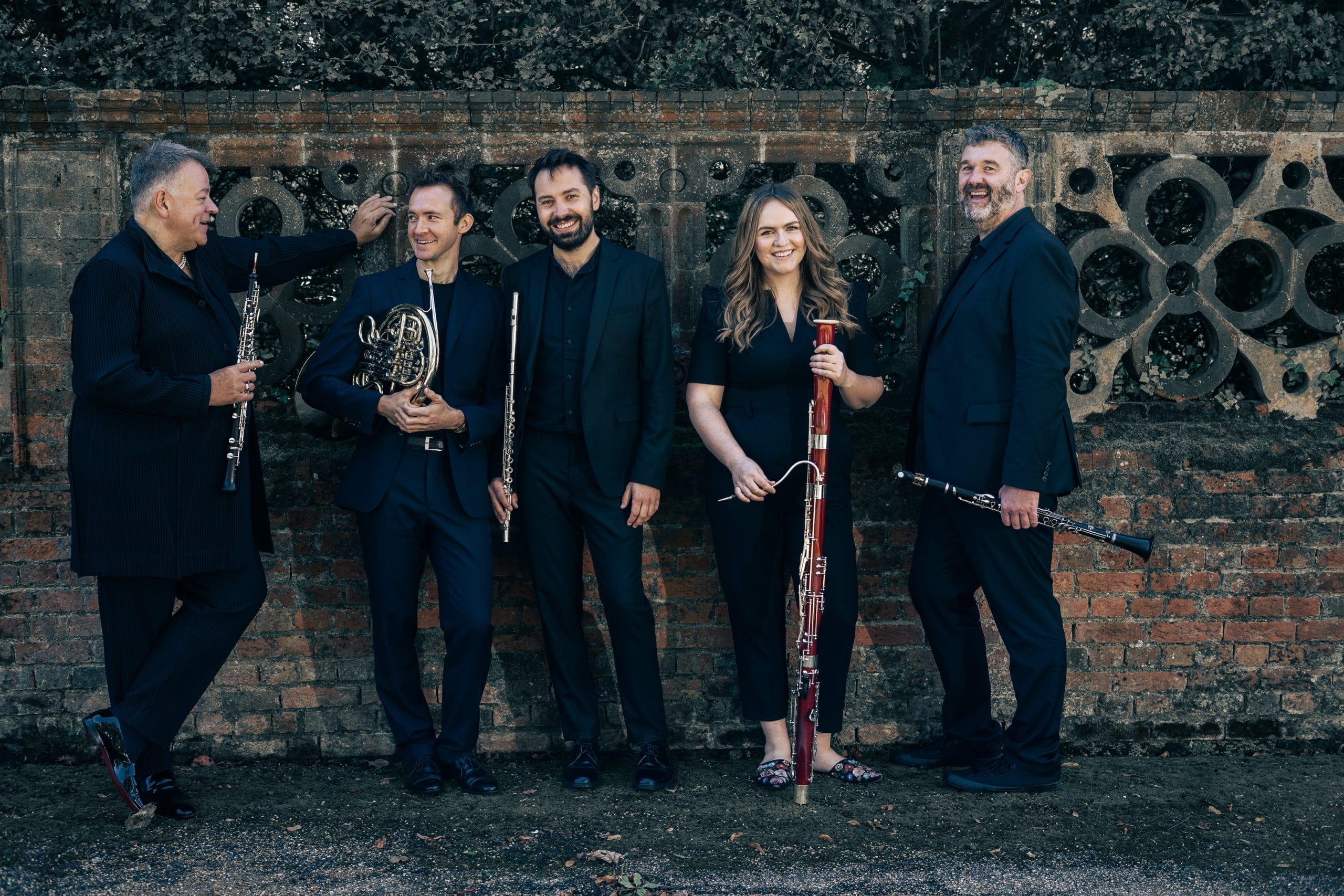 Five musicians, four men and one woman, all dressed in black are standing against an ornate stone wall holding their wind instruments.