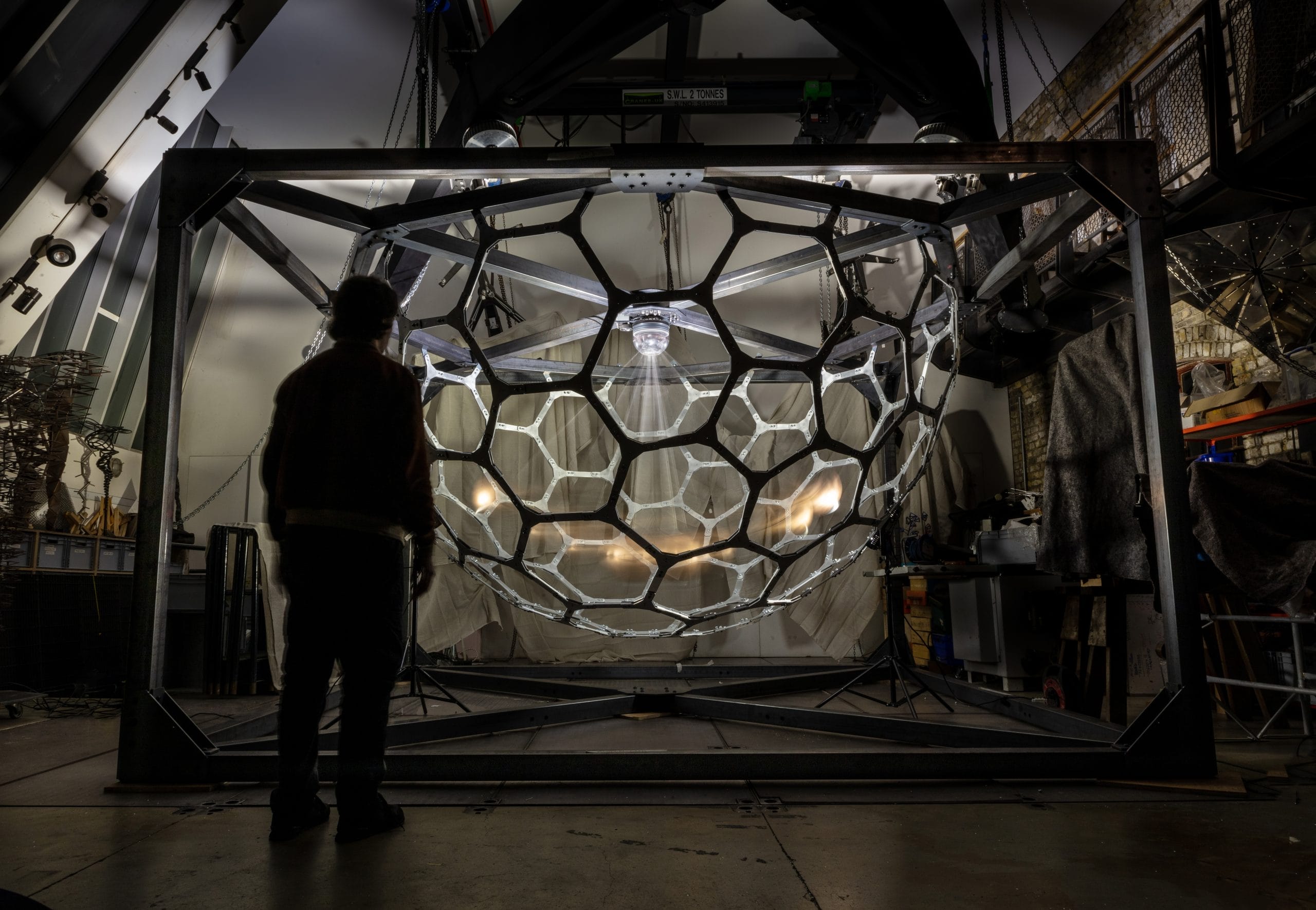 Artist Conrad Shawcross RA in his studio with Ring Down - a structural piece of artwork, a metal honeycomb dome with lights and a pendulum inside.