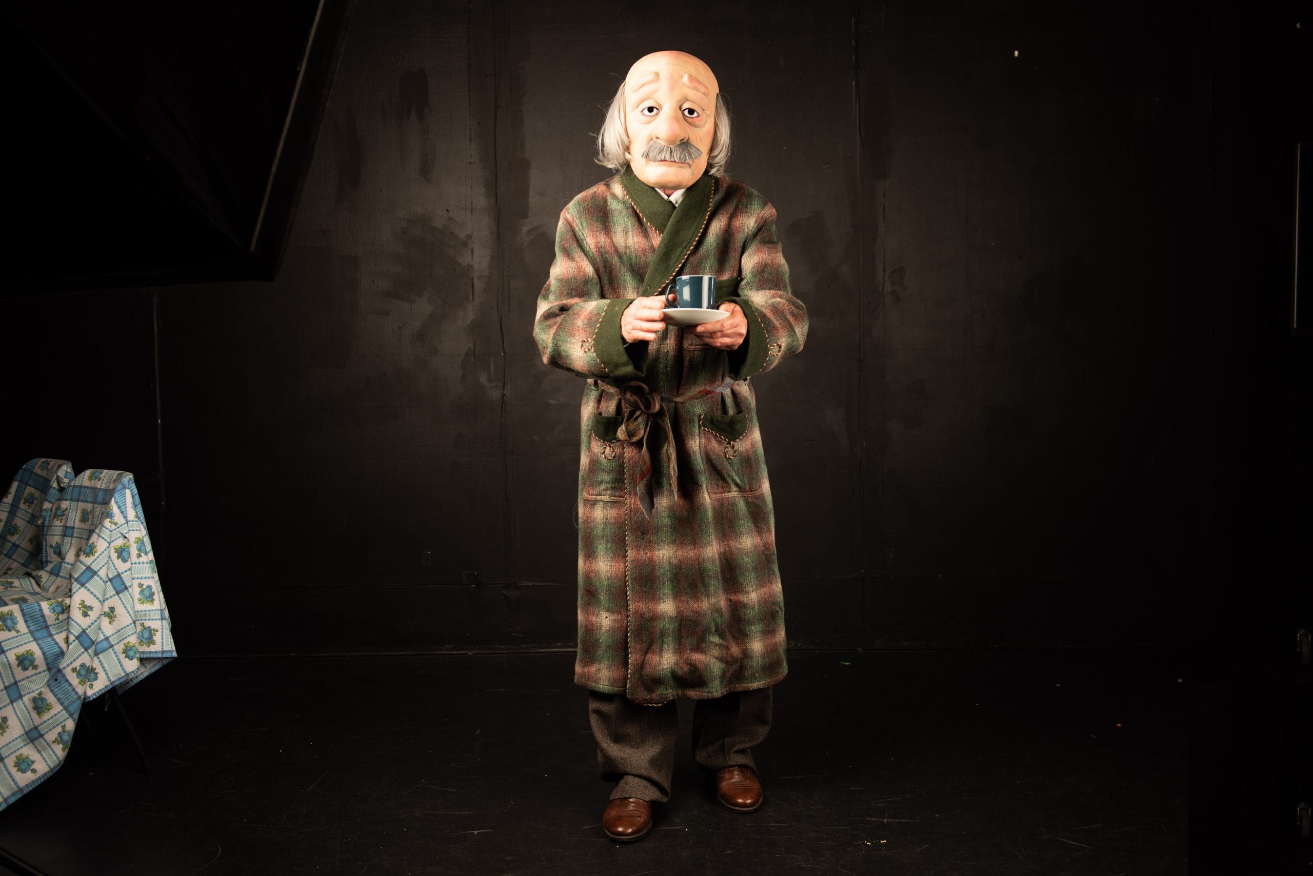 A masked old man with a cup of tea on a dark stage