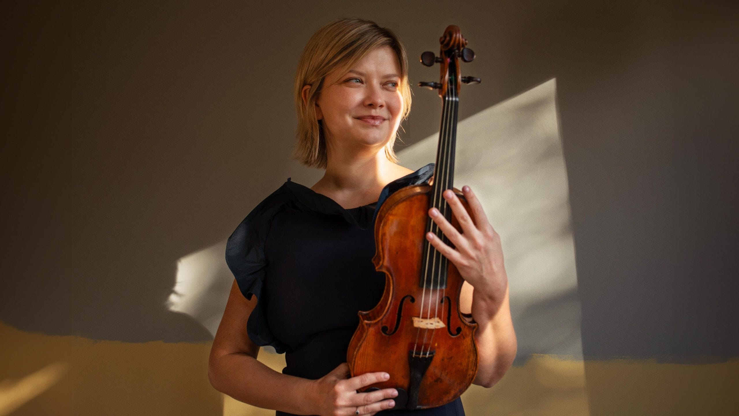 A woman with a short blond bob wearing a blue dress stands in the sunlight holding her violin.