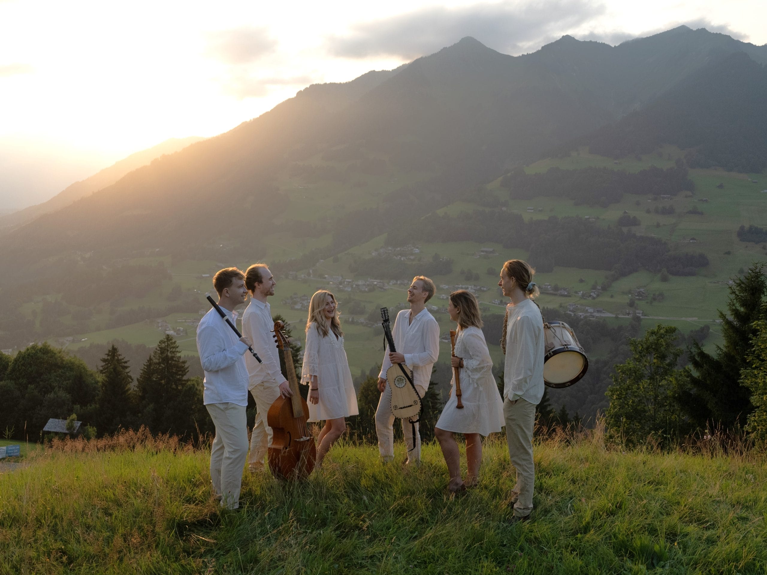 A group of six musicians, dressed in white stand on a green meadow with a mountain scape in the background.