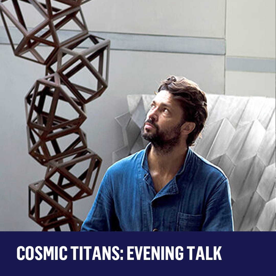 Artist Conrad Shawcross, a man in a blue shirt sits in a well lit space with a metal sculpture next to him.