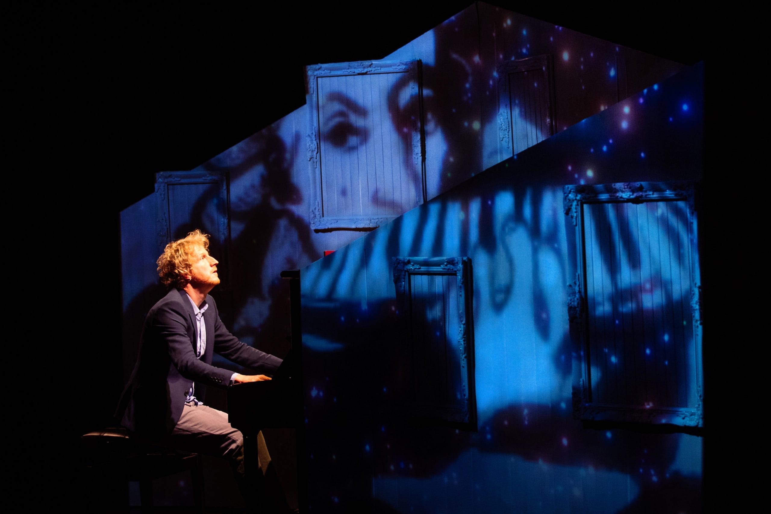 a blonde man playing a piano on stage with projections on the wall behind him