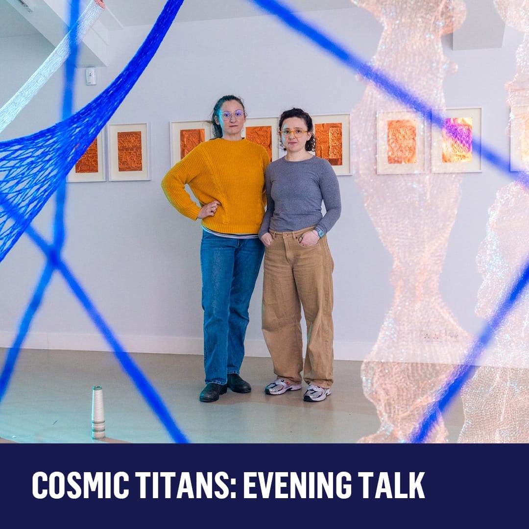 Two artists, one wearing a yellow jumper and jeans, the other a blue top and beige trousers, stand in front of framed copper engravings.