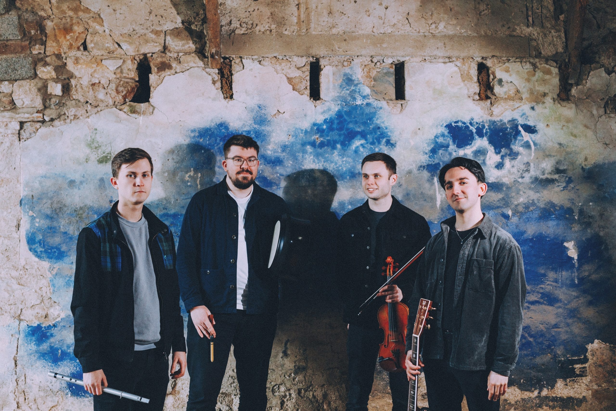 Four men stand in front of warehouse wall, painted blue and white, holding a selection of string instruments.