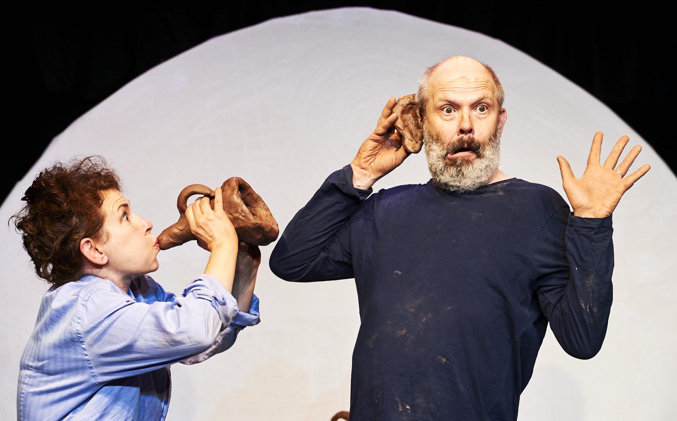 a woman blows into a trumpet made of clay into the ear of a surprised man