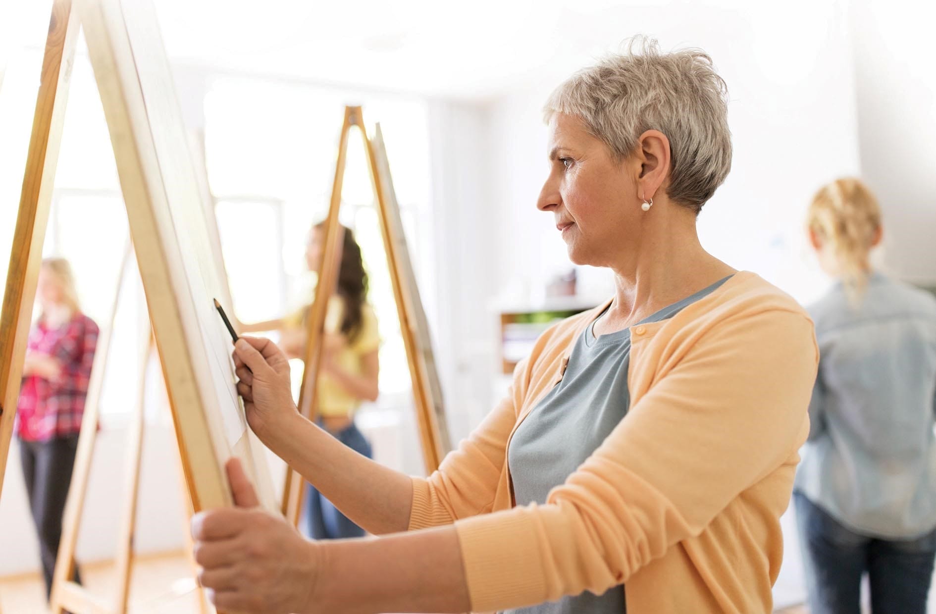 A woman with short grey hair, wearing a yellow cardigan stands at an easel drawing.