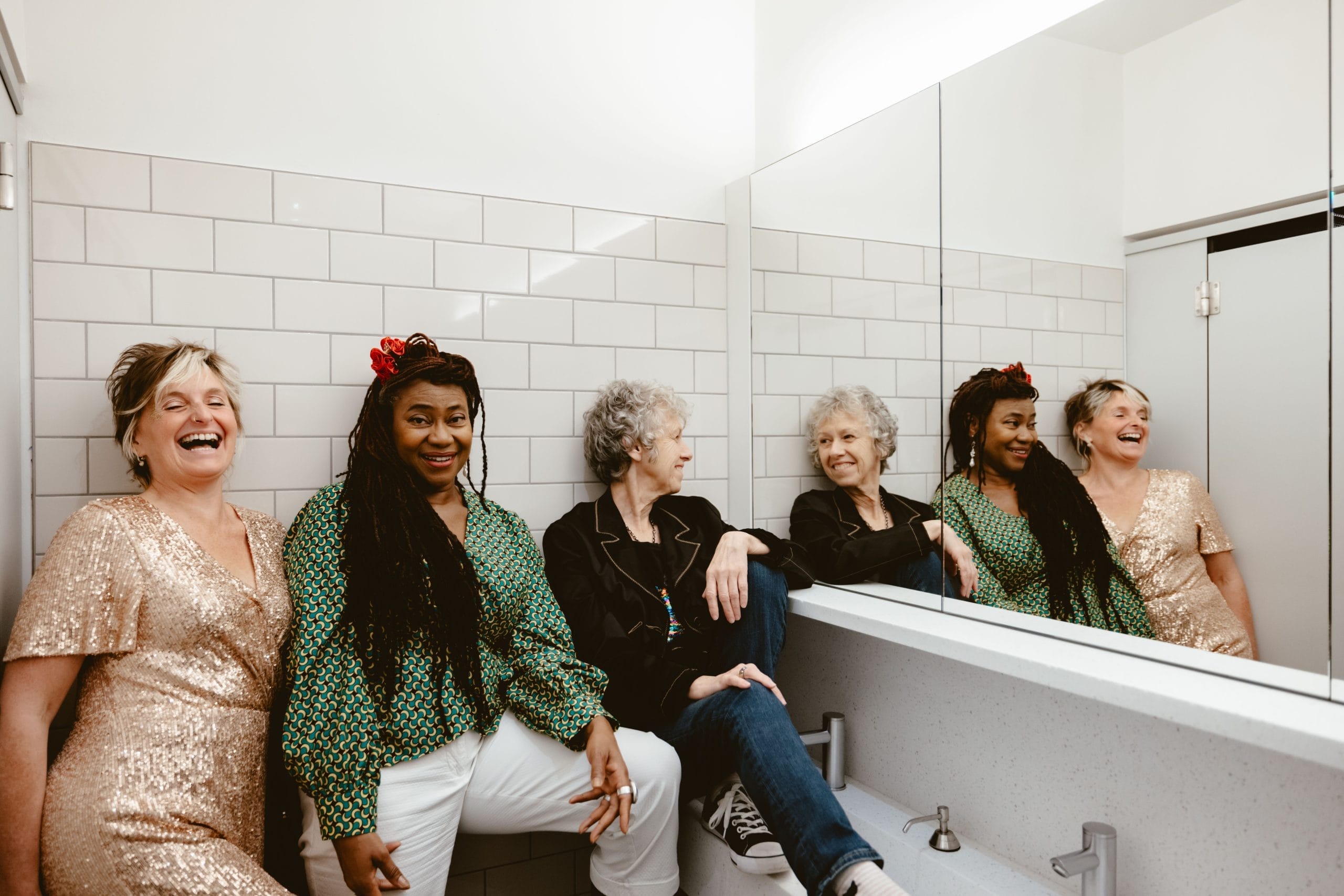 three women are smiling in a public toilet next to the sinks and mirrors