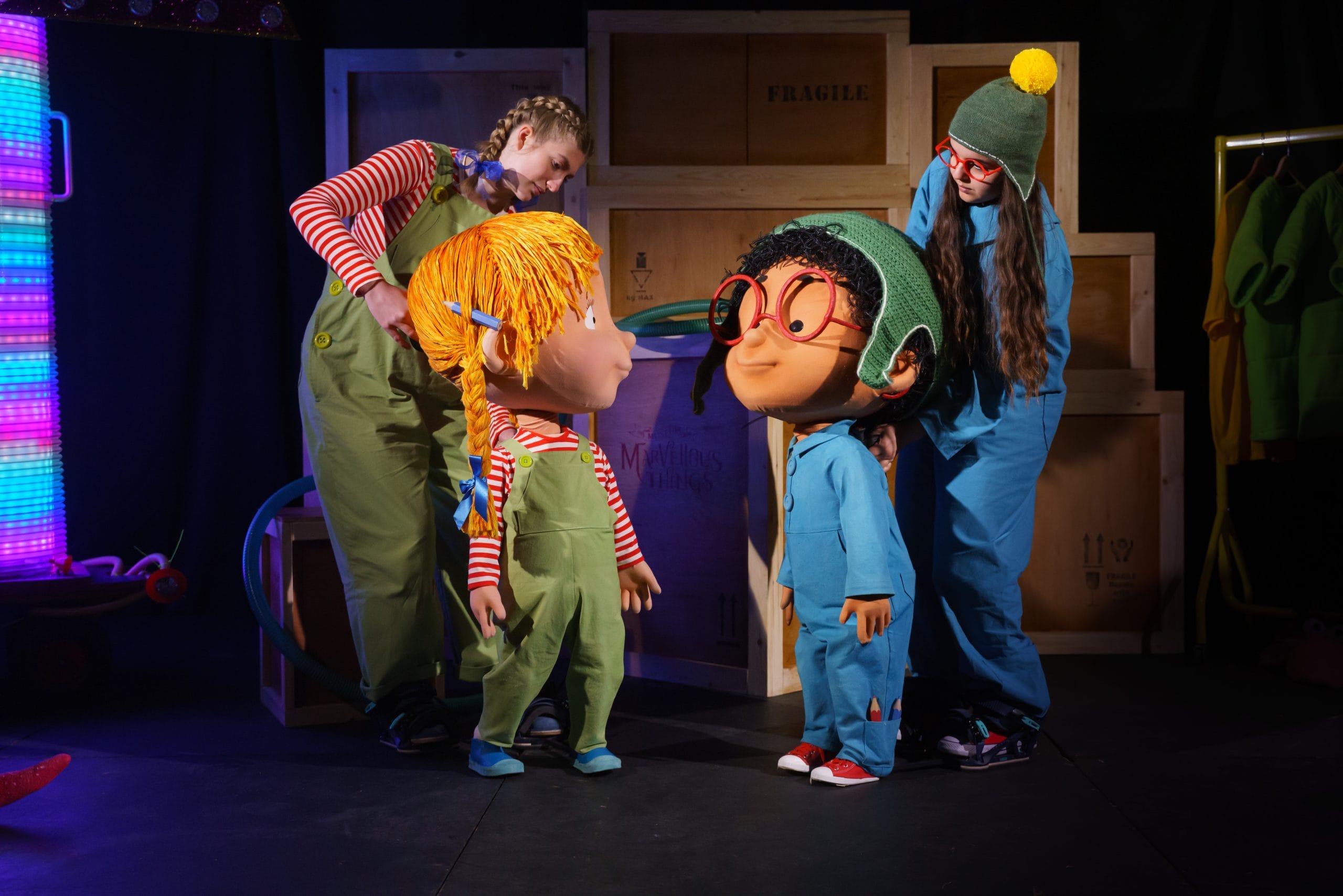 A pair of puppets stand on stage, one a girl with blond pigtails and green dungarees and a boy with blue overalls, red glasses and a green hat.