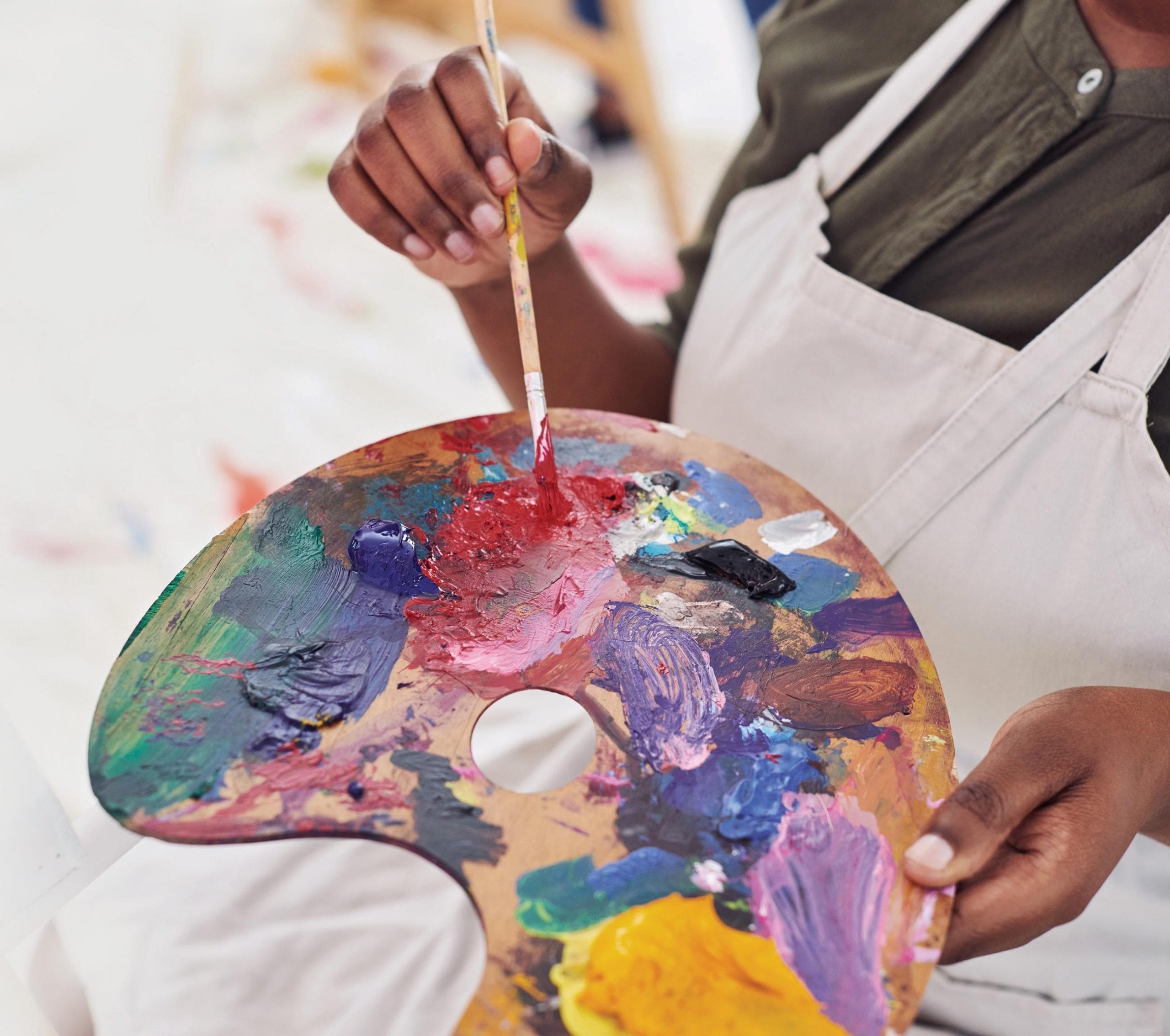 A paint palette loaded with various colours of paint, held by someone wearing a white apron.