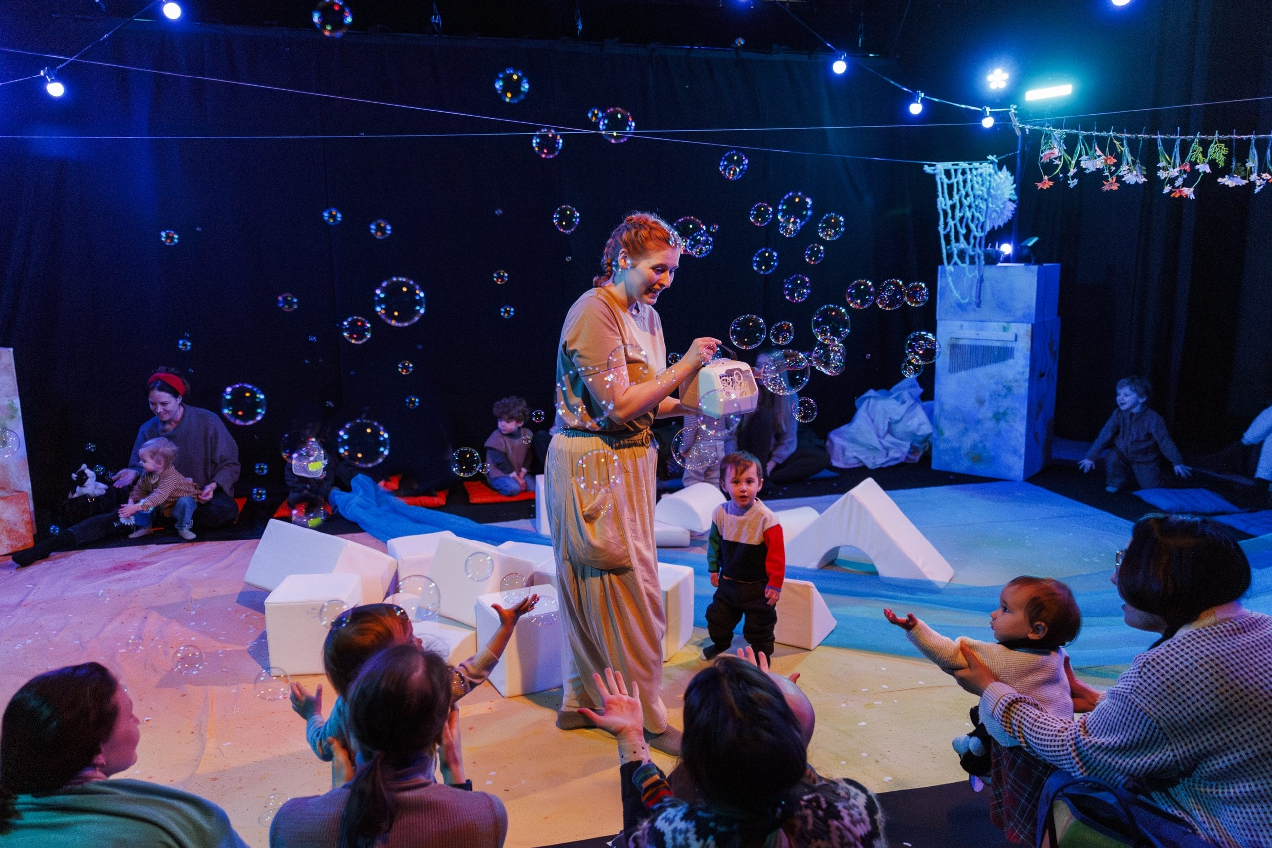 A lady stands surrounded by small children, holding a bubble machine, as bubbles fill the air.