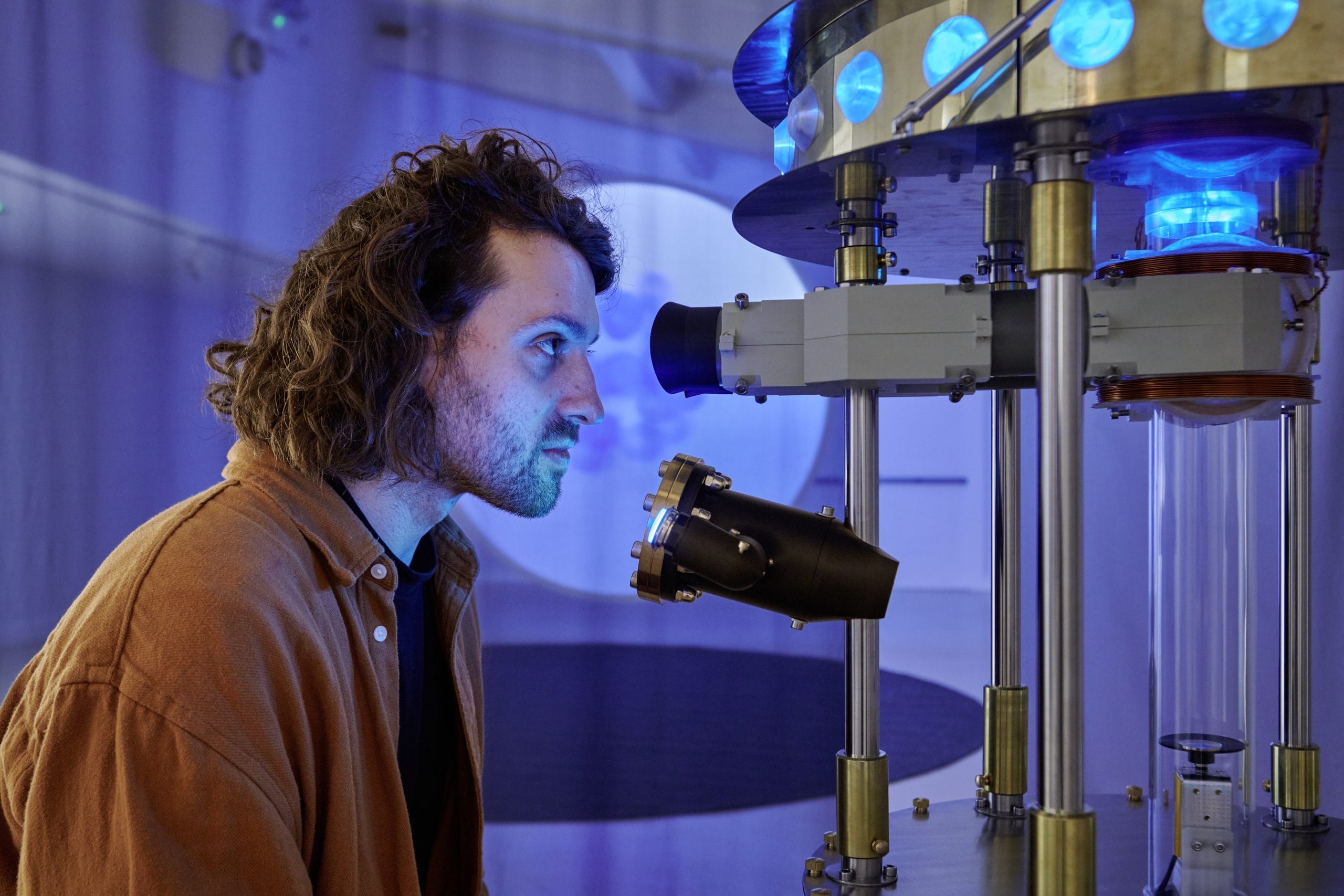 A man, with dark hair to his chin wearing a brown shirt looks in to a quantum computer.