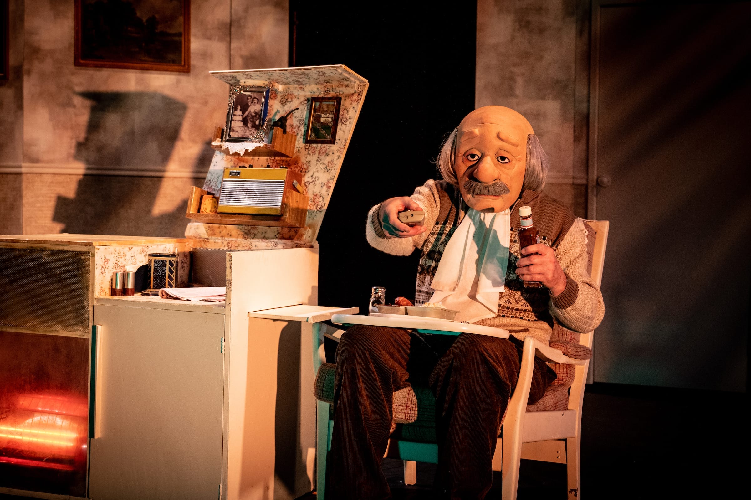 A photo of a masked old man turning the TV on with his dinner in front of him on stage.