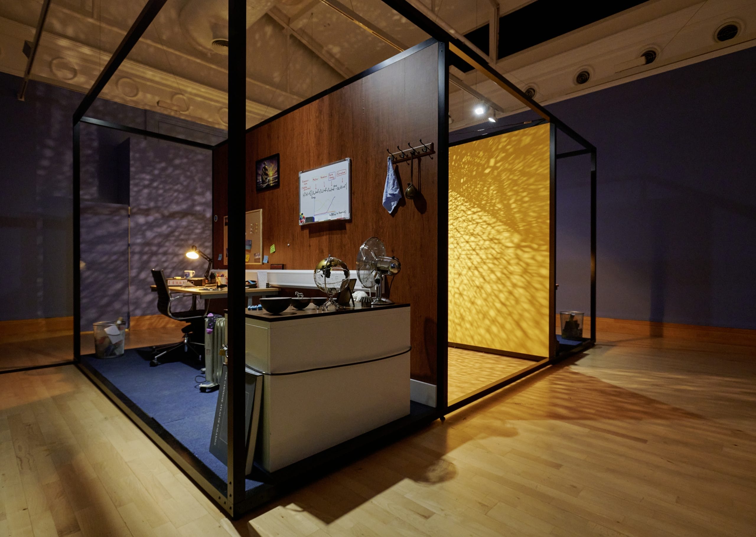 A sculptural installation of an office space, with a yellow wall behind with intricate shadows being cast.