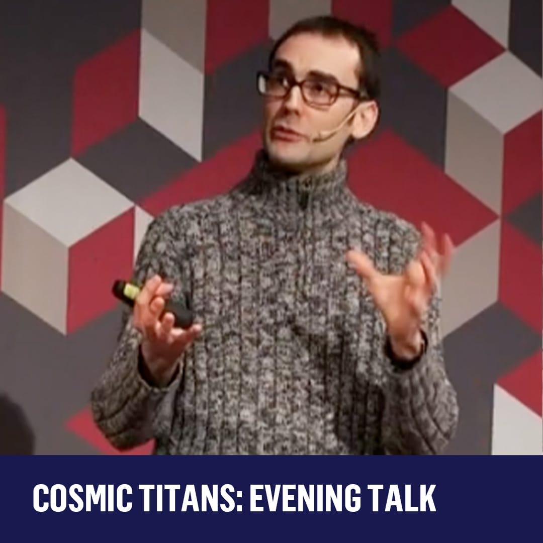 Professor Andrew Pontzen, wearing a grey roll neck jumper, glasses and short dark hair stands in front of a geometric print wall.