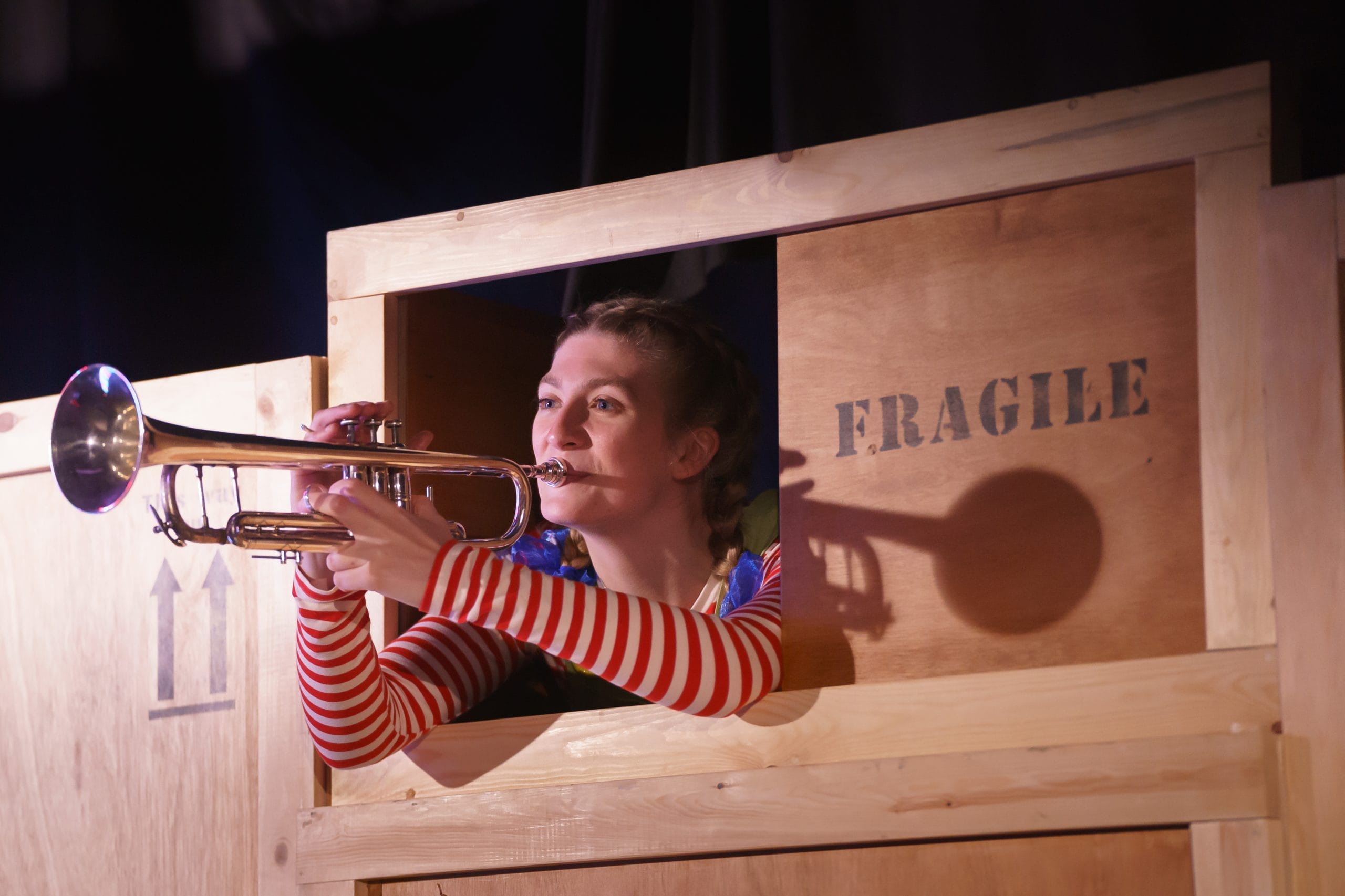 A photo of an actor playing the trumpet sitting in a box labelled FRAGILE in The Museum of Marvellous Things