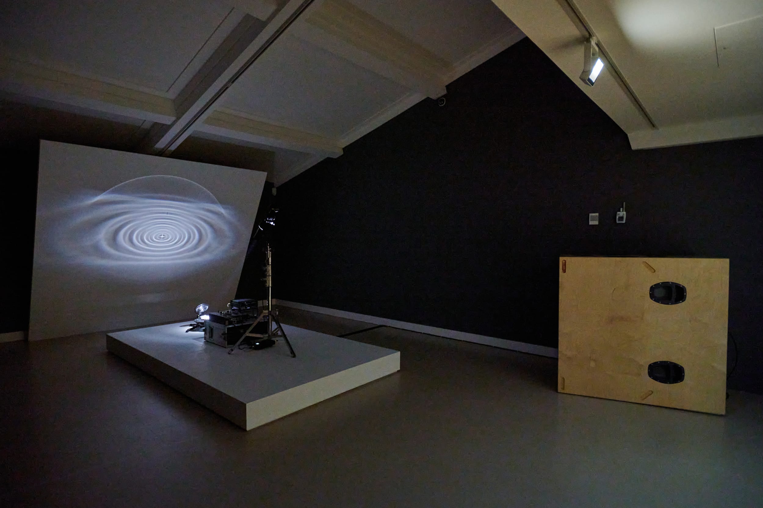 A dark gallery room features a large screen, projector and speaker.