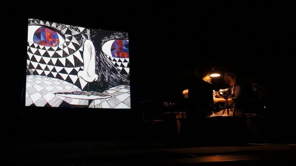 on a dark stage, there is a projected drawn face as well as people under light who are holding paper cut outs to project