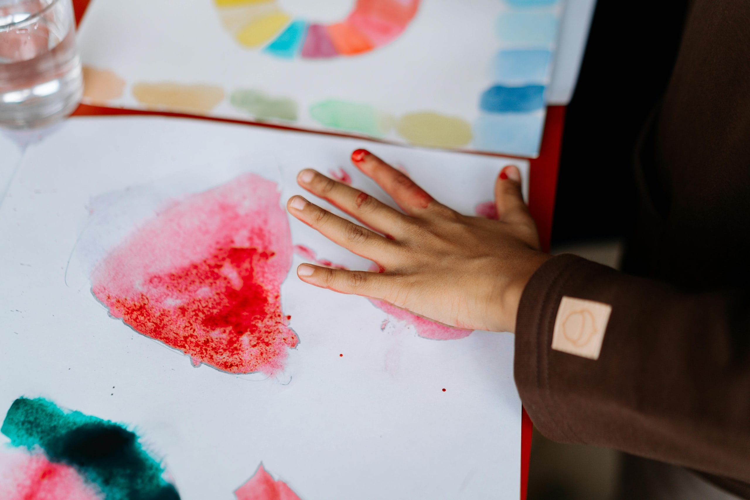 White paper, with painted shapes covers a table, a child presses their hand, covered in red paint on to the paper