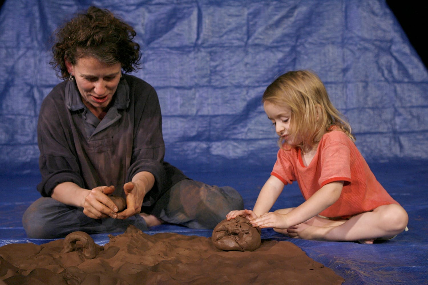 a little girl in a pink top sits next to a woman and they both play with clay