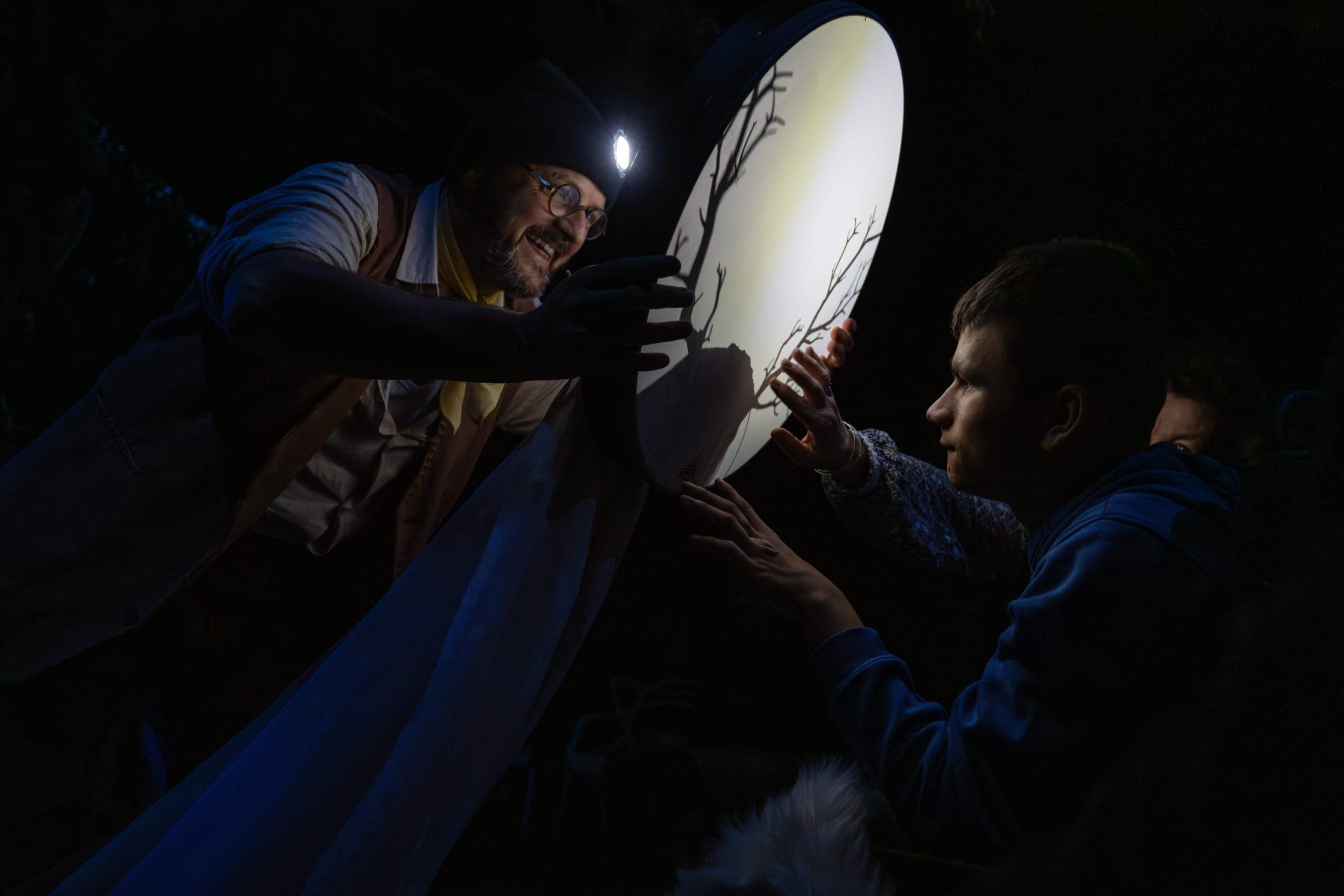 An actor wearing glasses is holding a white canvas stretched on a circle to make the shadow of a stag that is viewed by an audience member