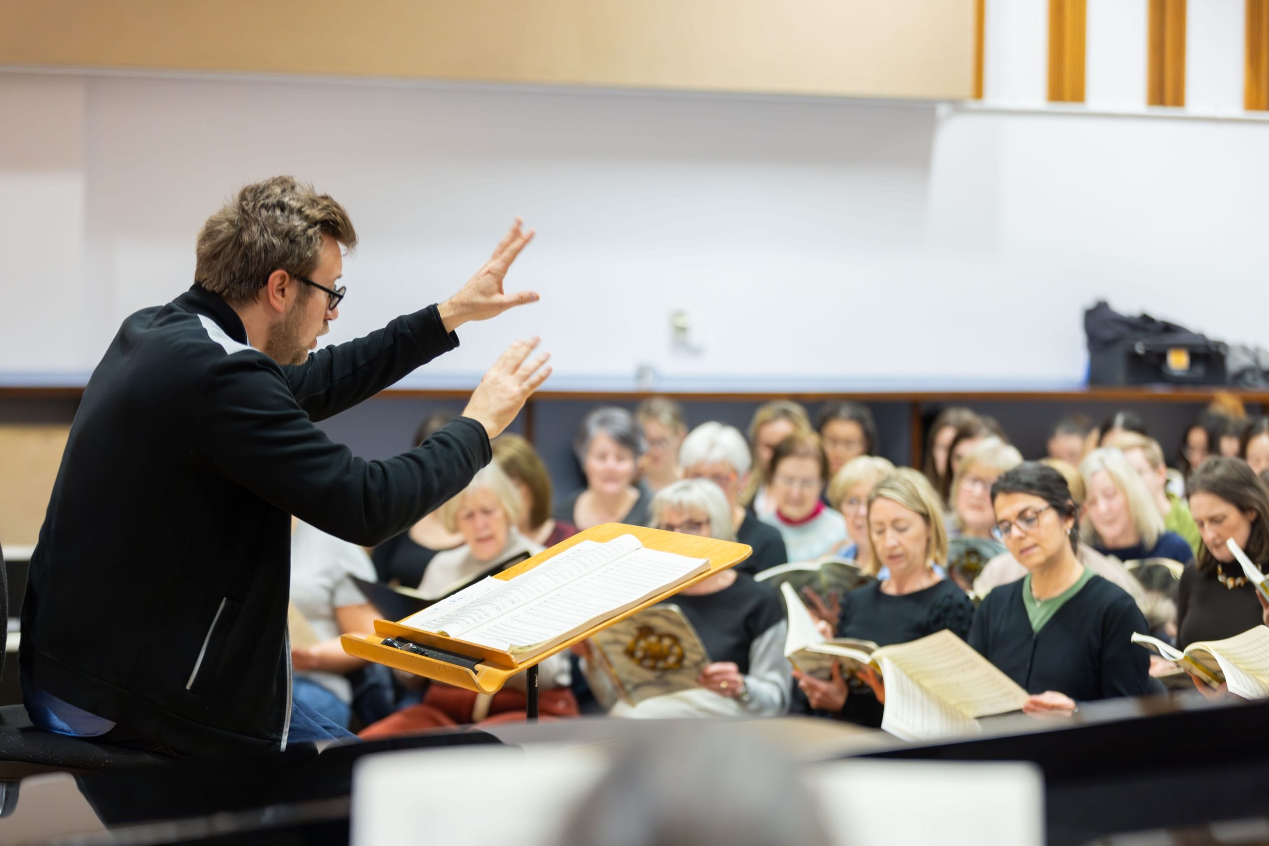 a male conductor instructing seated singers in a rehearsal hall