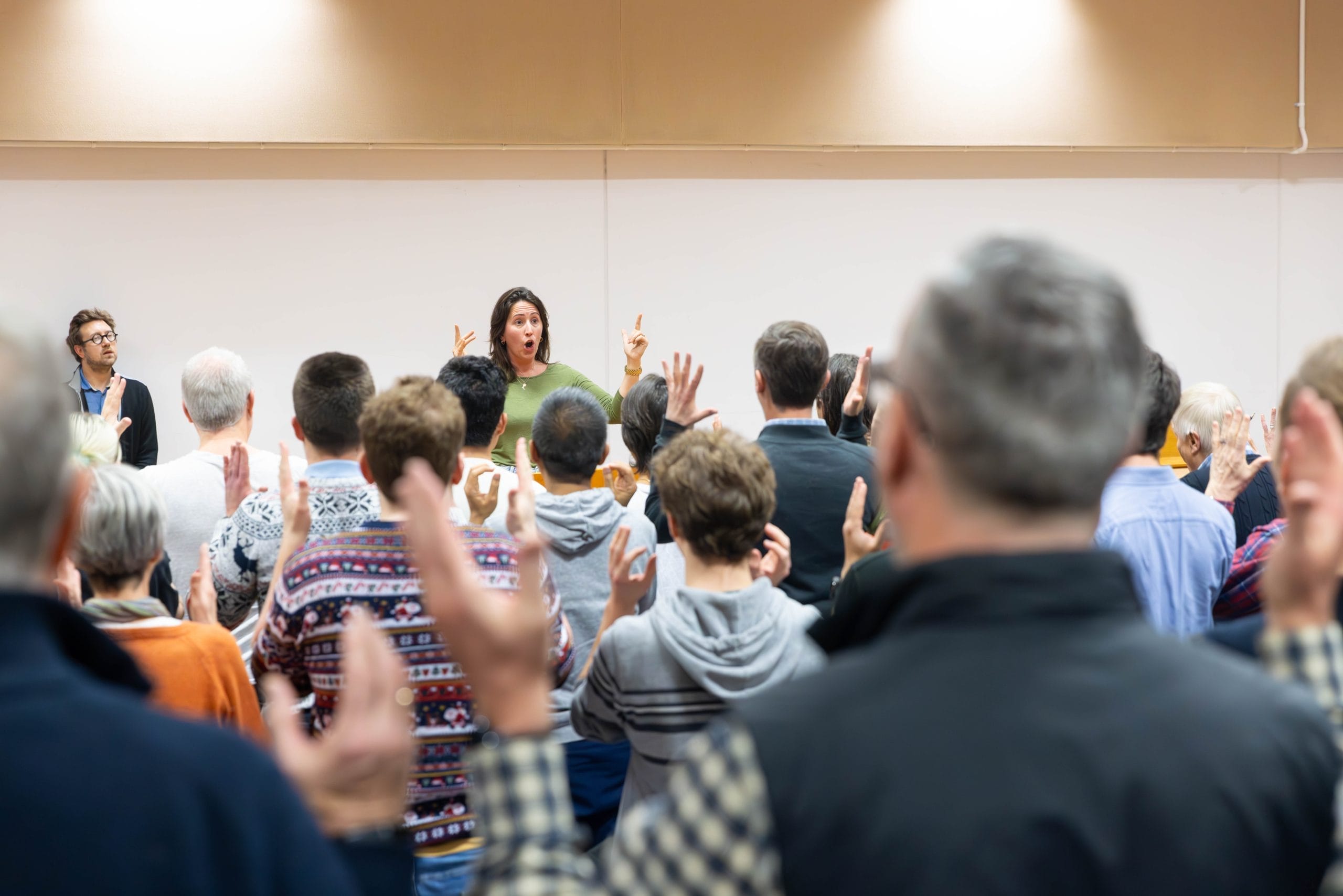 a group of people photographed from the back singing with their conductor visible in the front