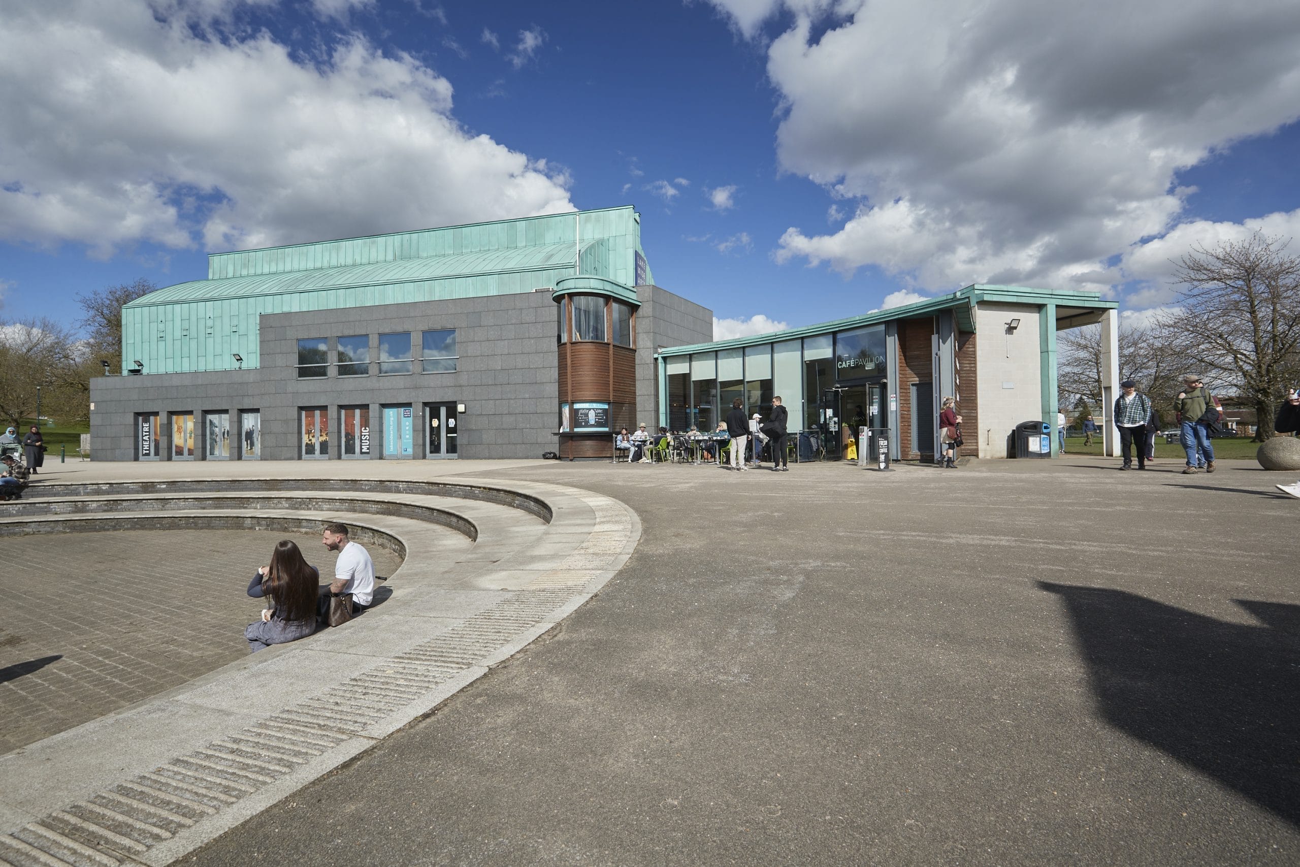 A photo of Lakeside Arts, a grey building with a green roof, with people enjoying the sunshine.