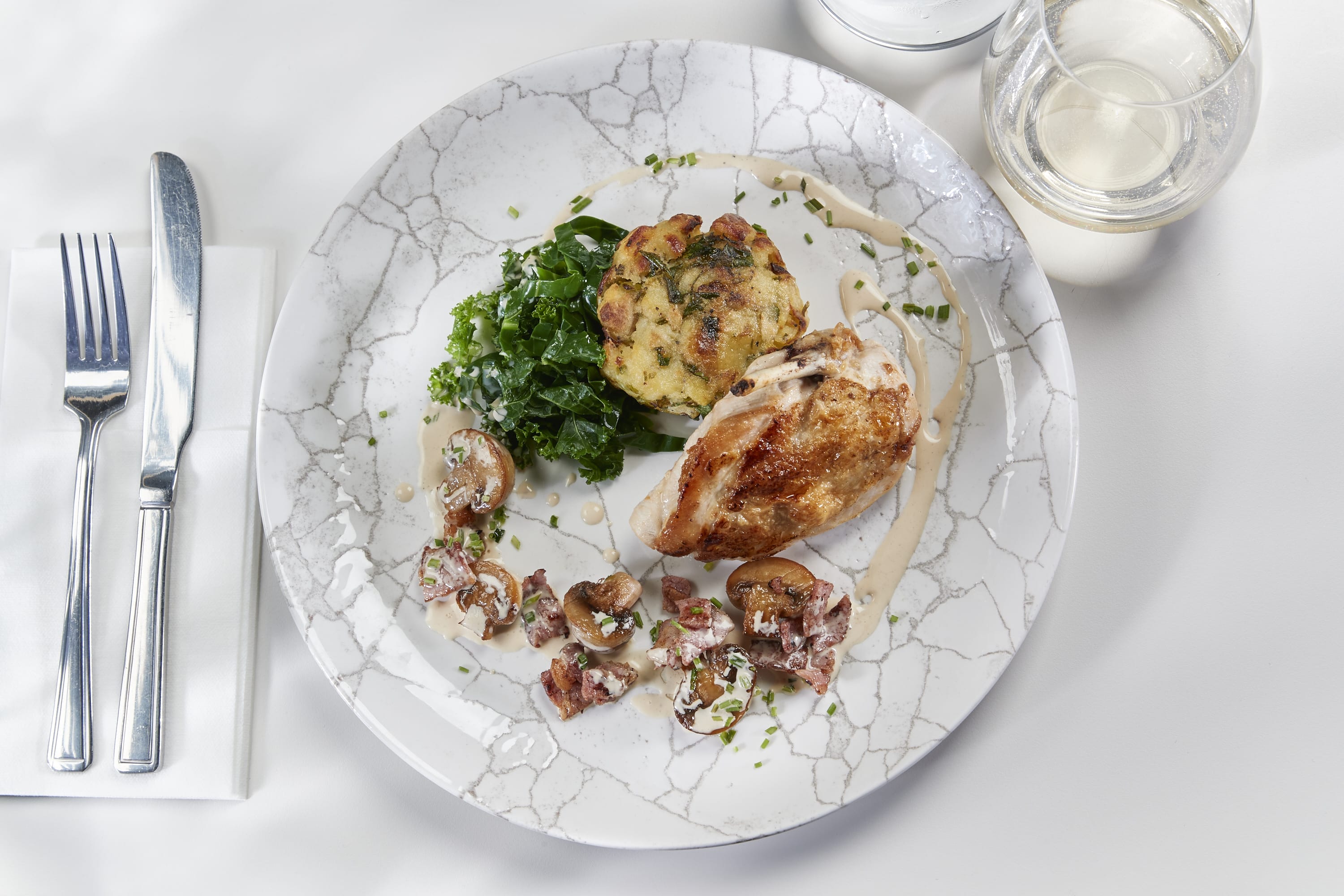 A white plate with crackled design holds a roast chicken breast, herb potatoe cake, wilted spinach, wild mushroom and bacon cream.