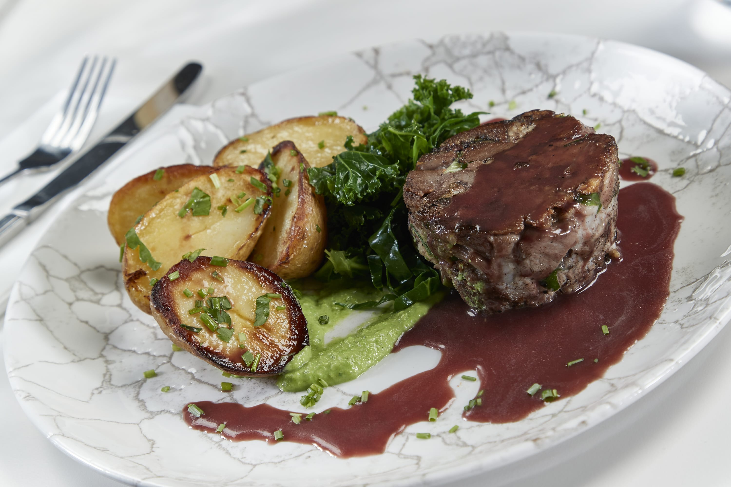 A white plate with a crackled design holds rolled lamb shoulder, spring greens, roasted baby potatoes, pea puree and a Red Wine Jus