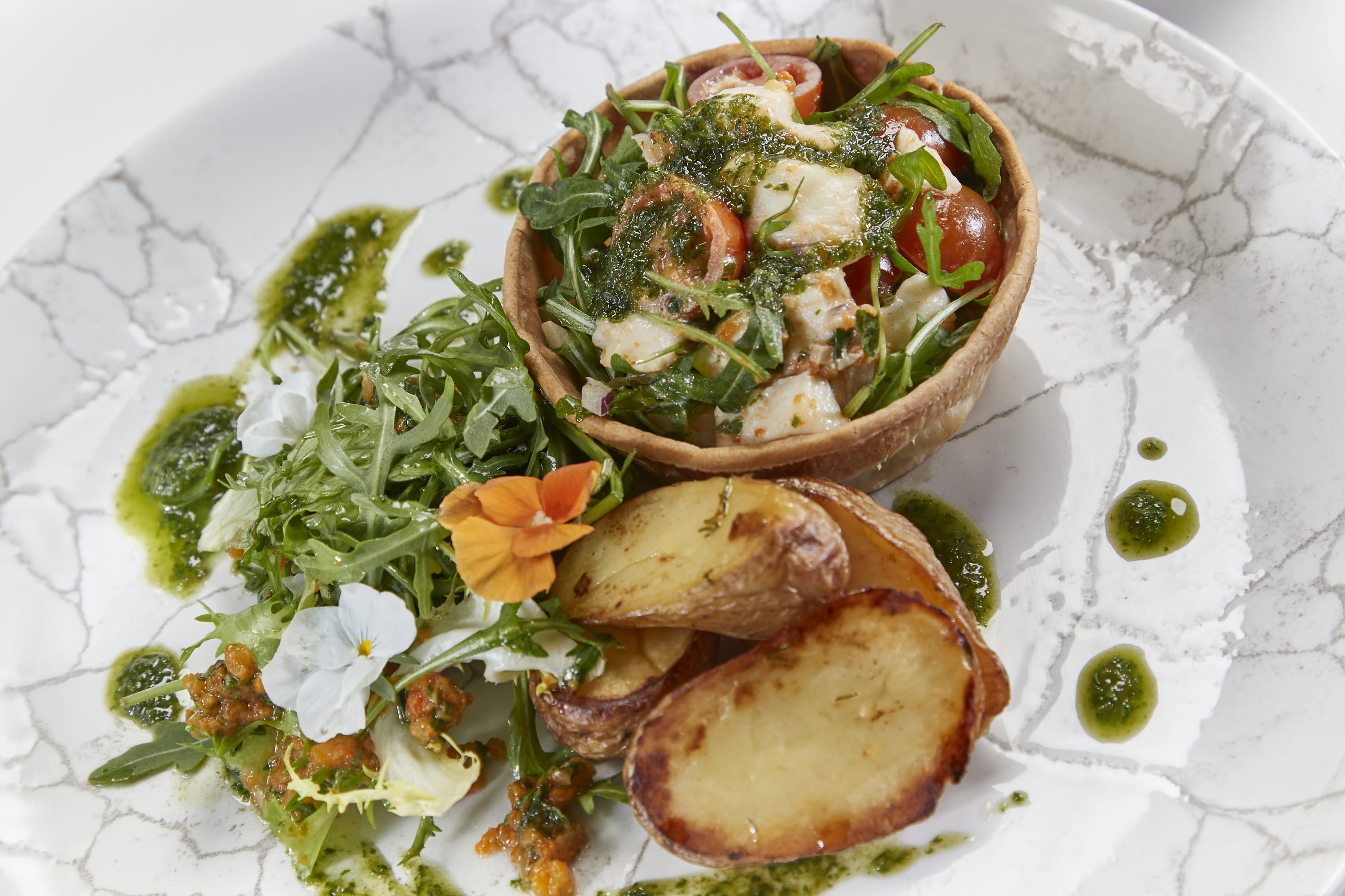 A white plate with crackled design, holds new potatoes, a pastry case, rocket and dressing.