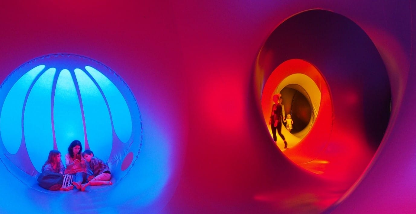 a landscape photo of the interior of the luminarium filled with blue and red light, with a family enjoying it while sitting