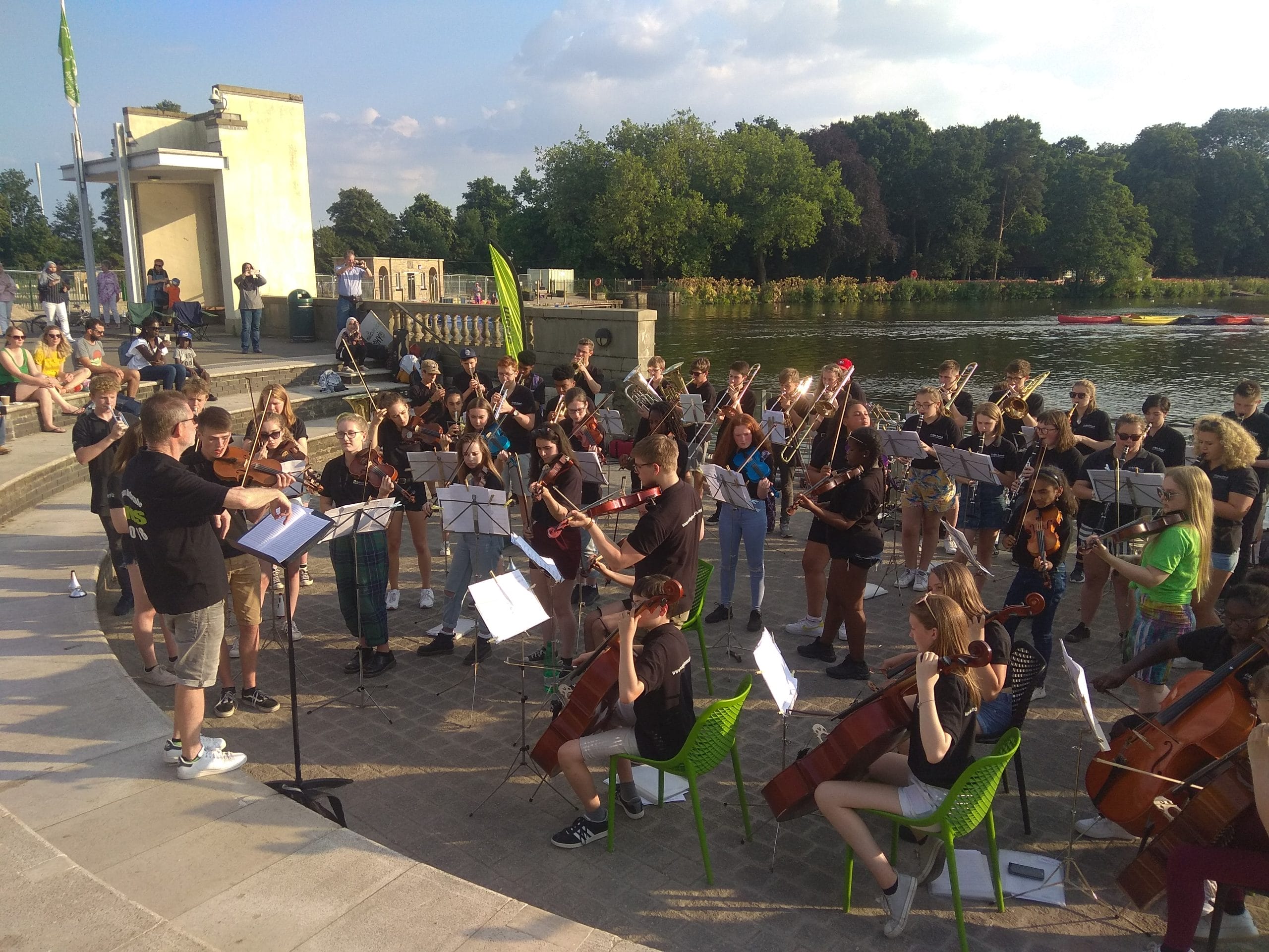 A large group of student musicians play in an amphitheatre by a lake.