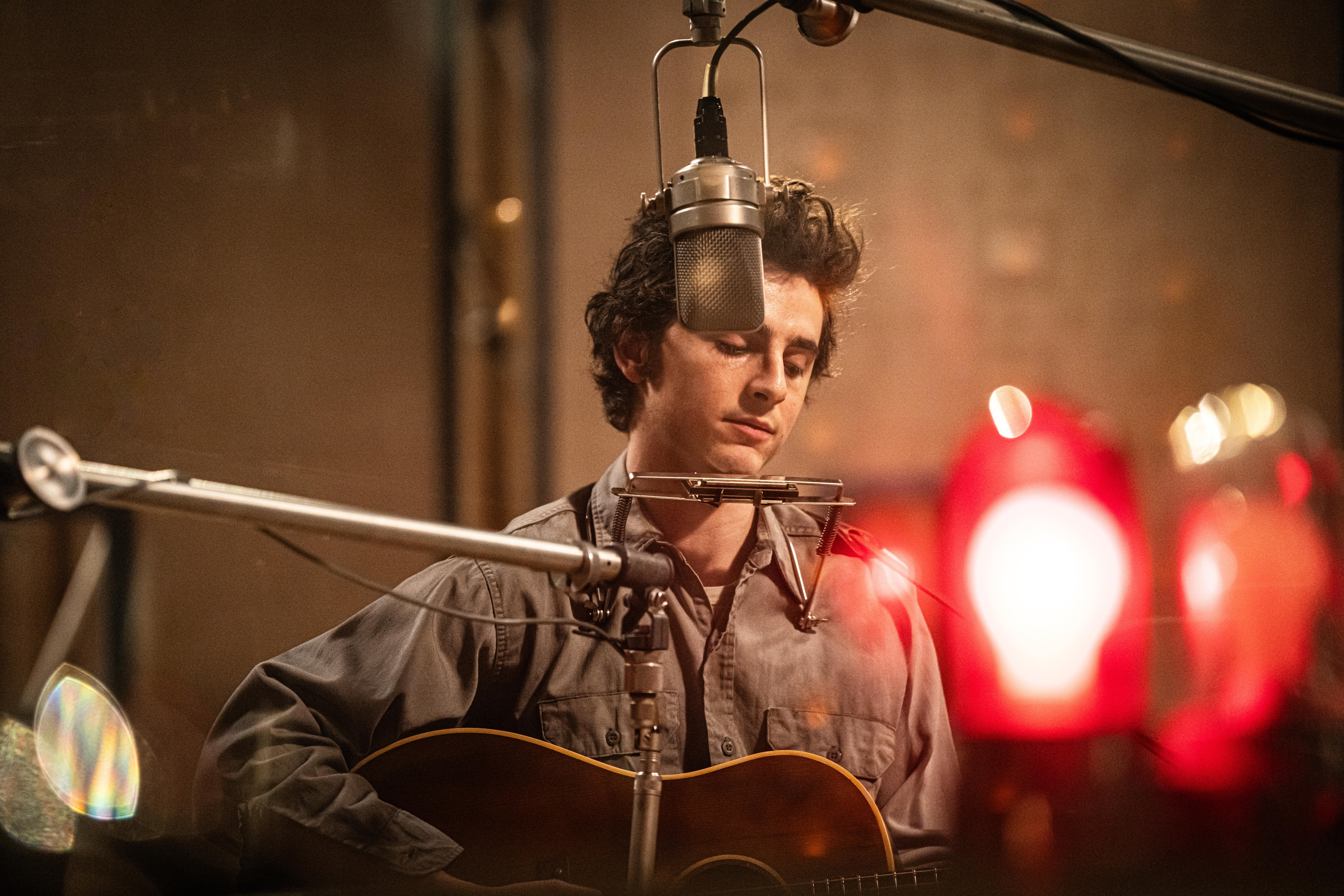 A young man sits in a recording studio, behind a microphone, holding a guitar.