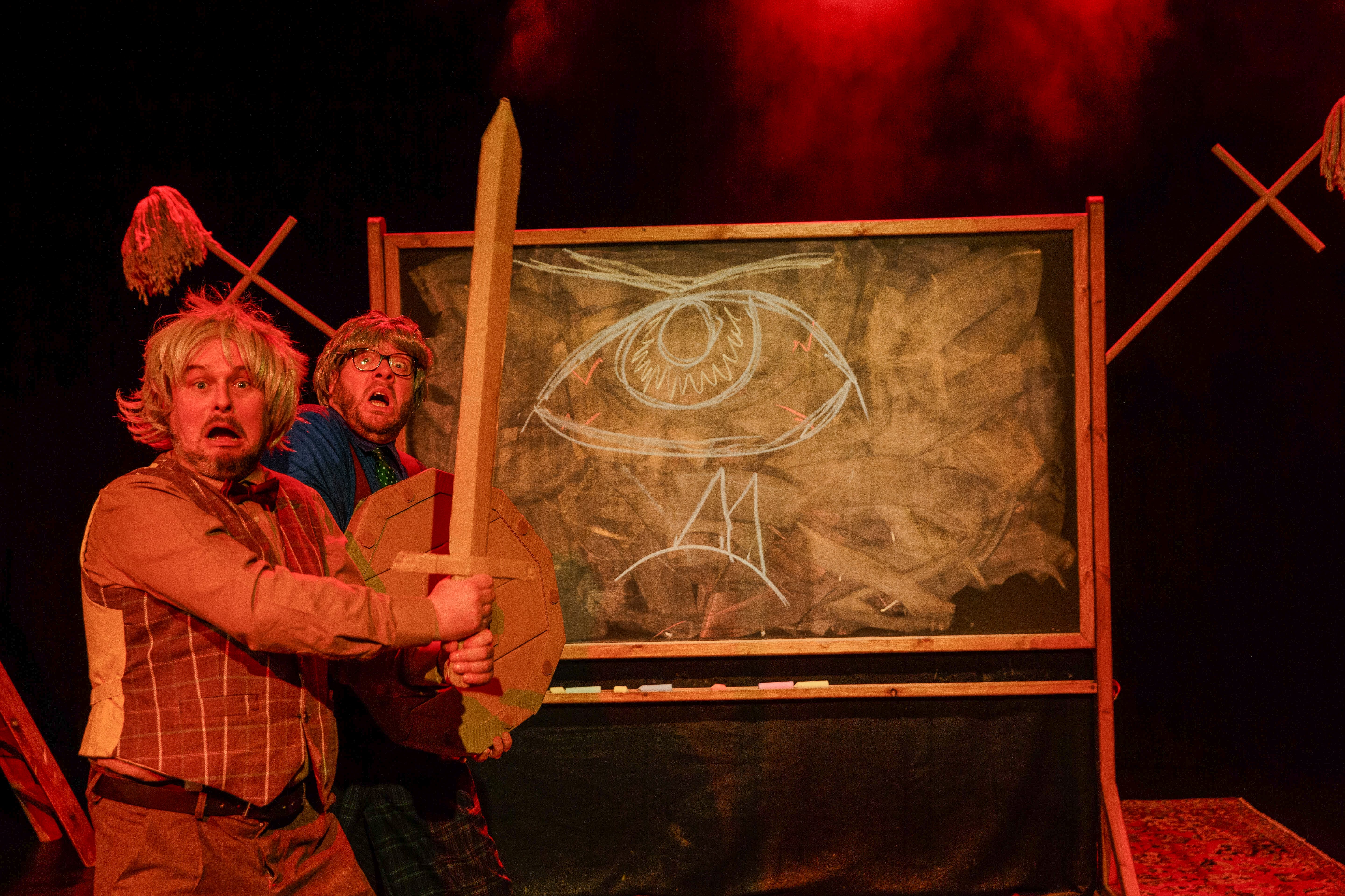 Two actors dressed as mad history professors wield a cardboard sword, in front of a blackboard.