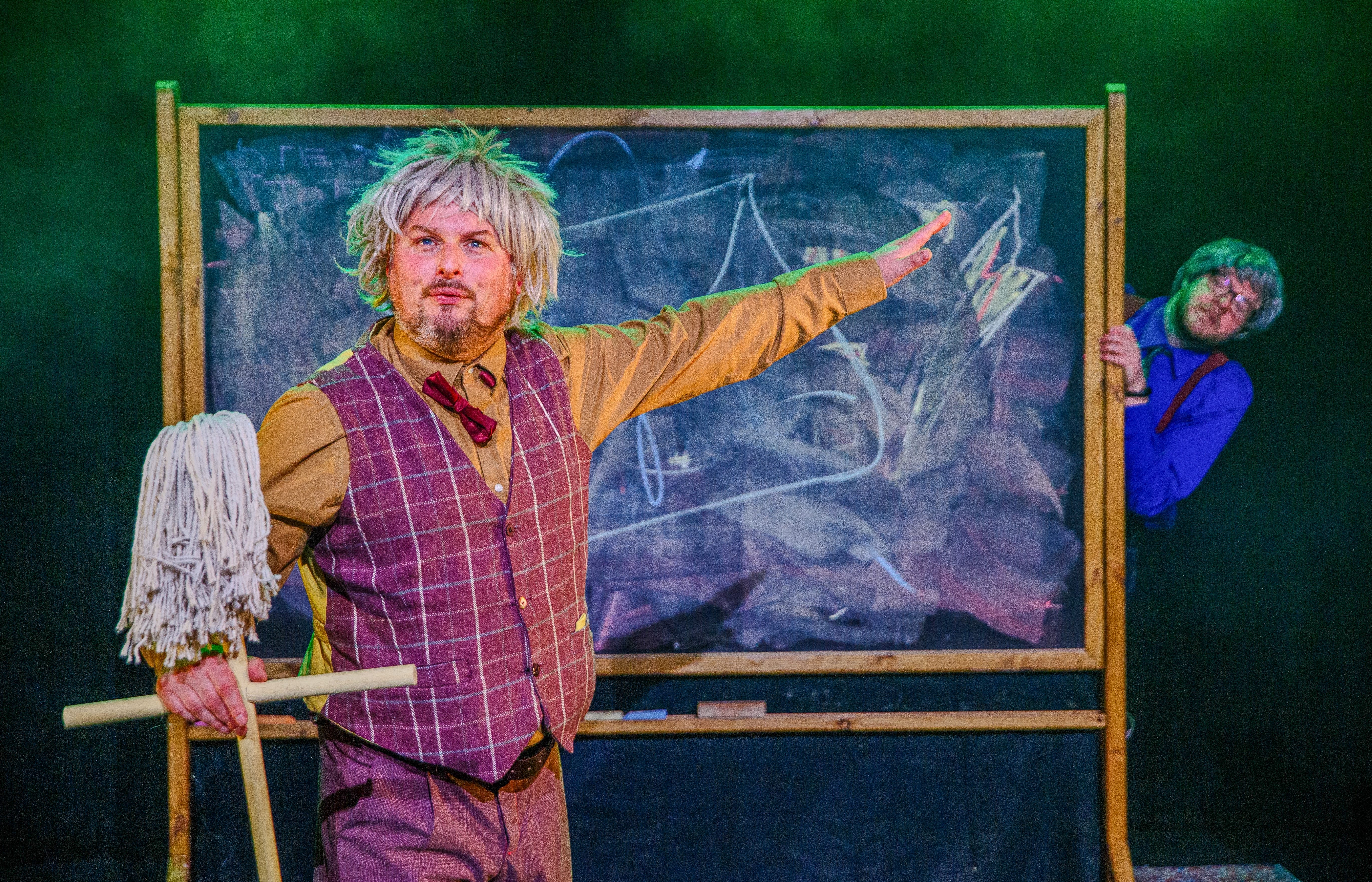 An actor dressed as a mad professor stands in front of a chalk board.