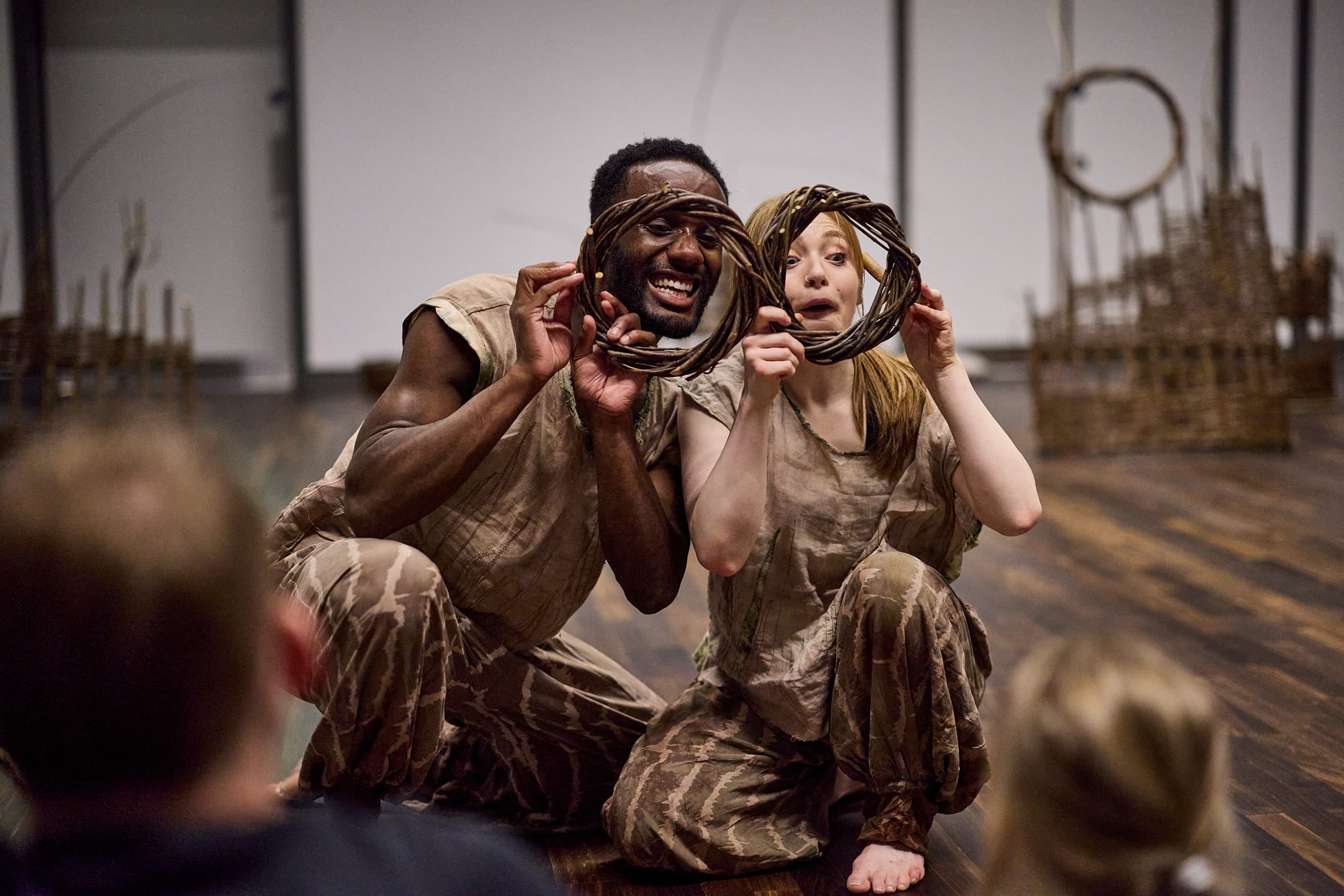 Two dancers, dressed in green peer through willow rings.