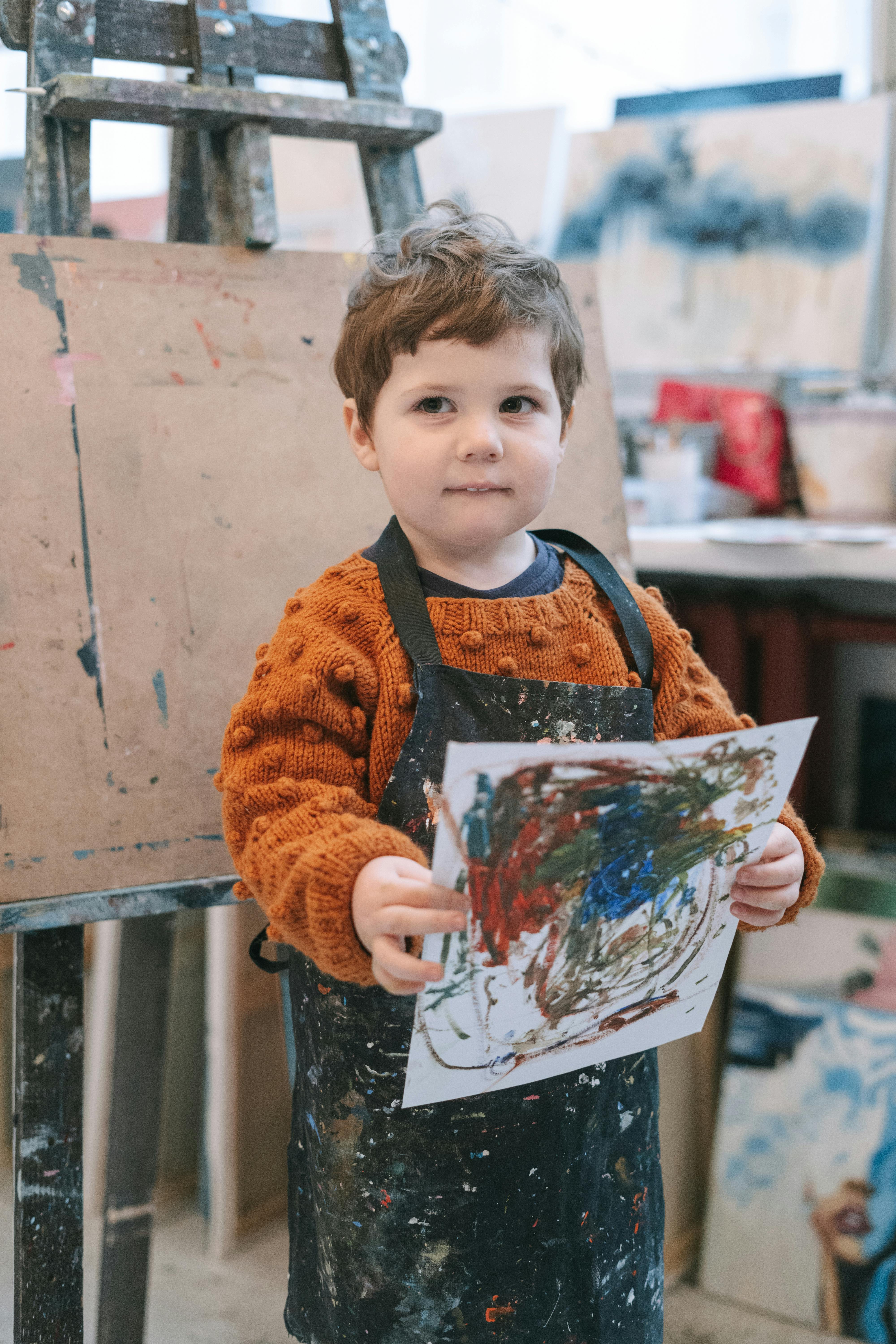 A small boy wearing an orange jumper and dark apron holds a messy painted picture.